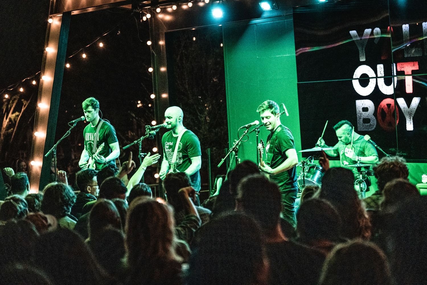Rock band performing on stage with Out Boy sign and string lights in background.