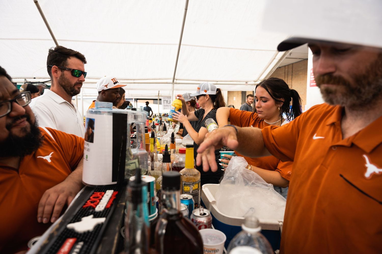 Crowd gathers around a bar serving drinks under a white tent.