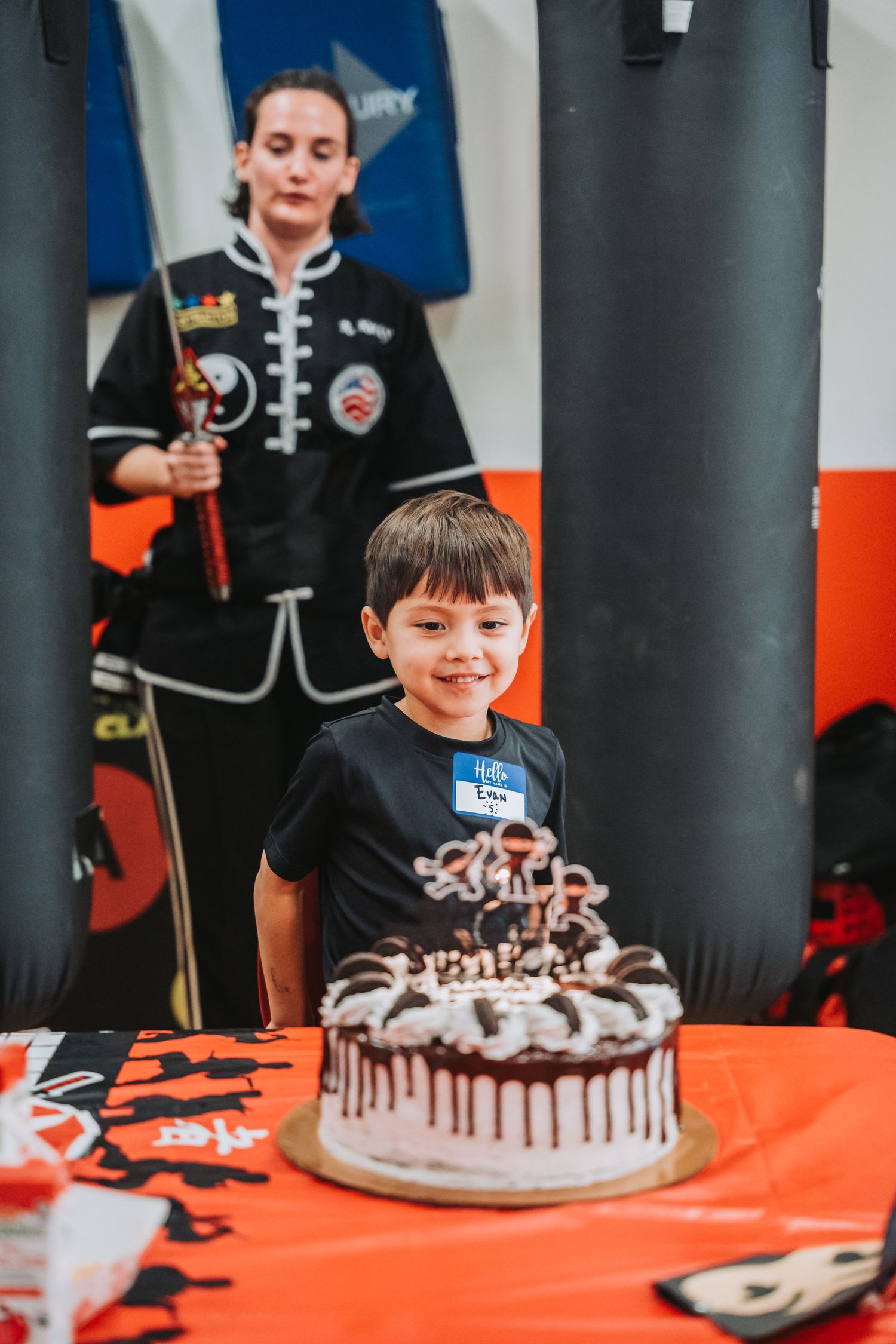 Young student stands proudly next to decorated birthday cake in martial arts studio.