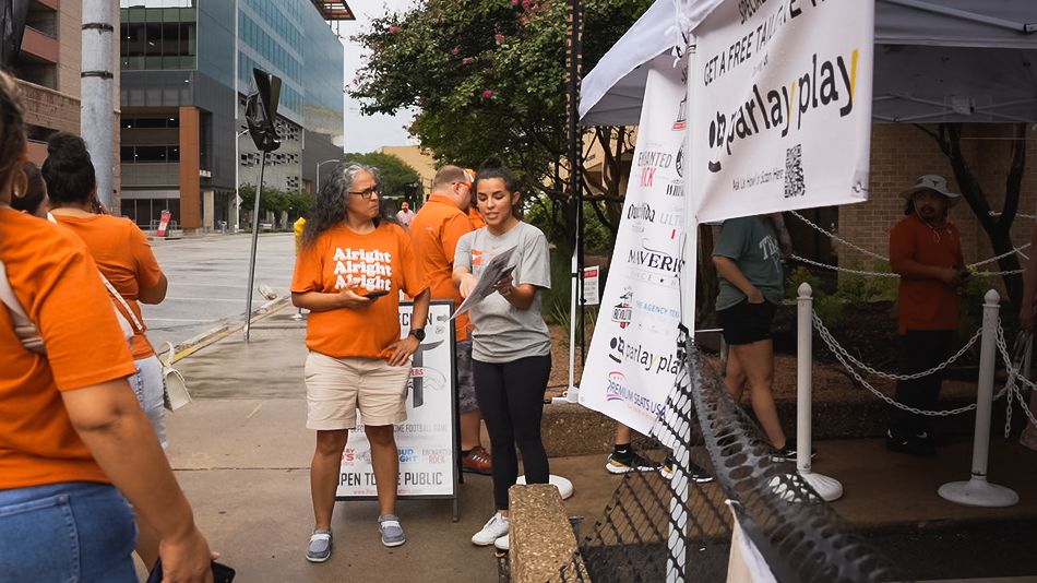 People in orange shirts gather at an outdoor event booth on a sunny day.