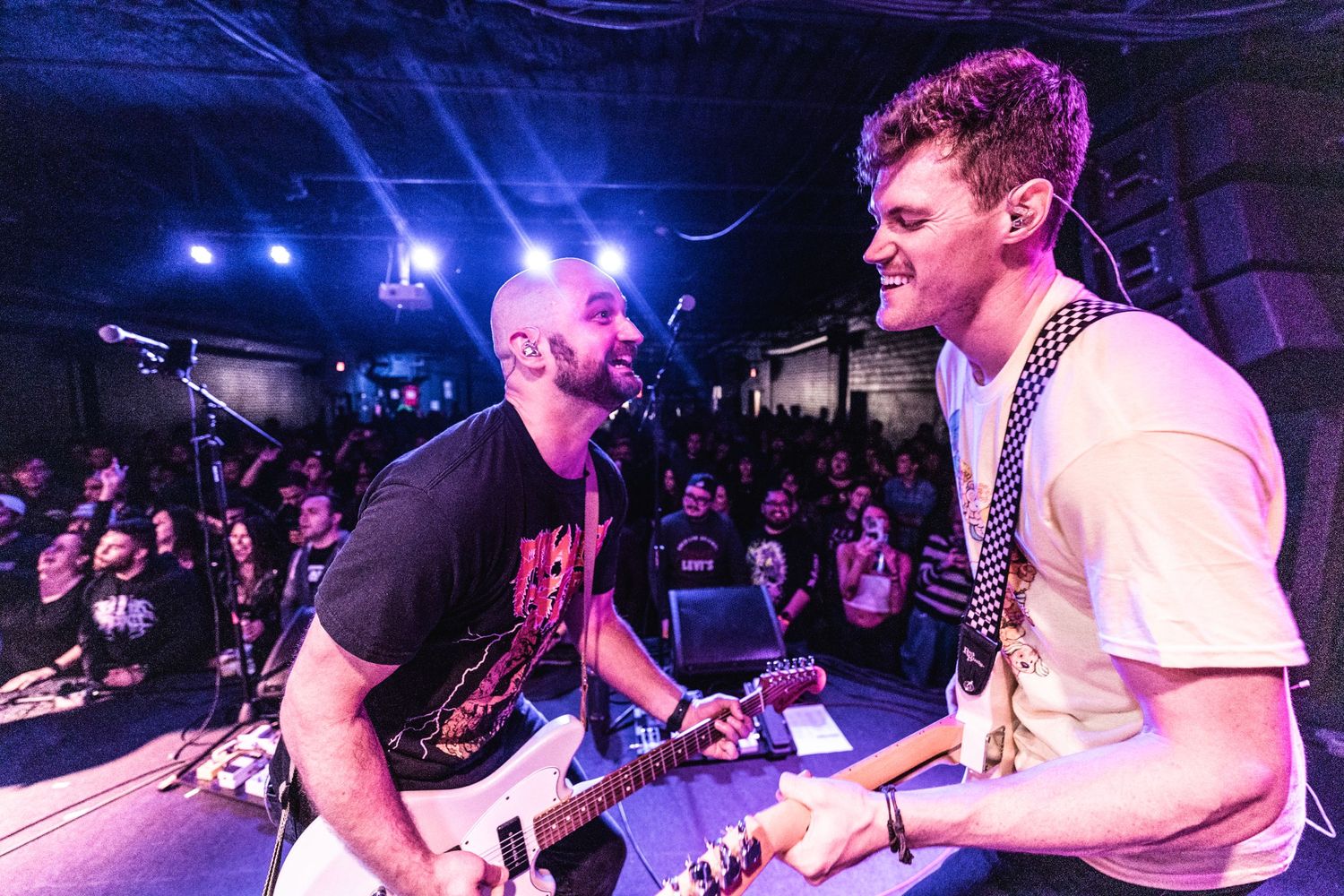 Musicians perform passionately on stage with blue lighting and crowd visible in background.