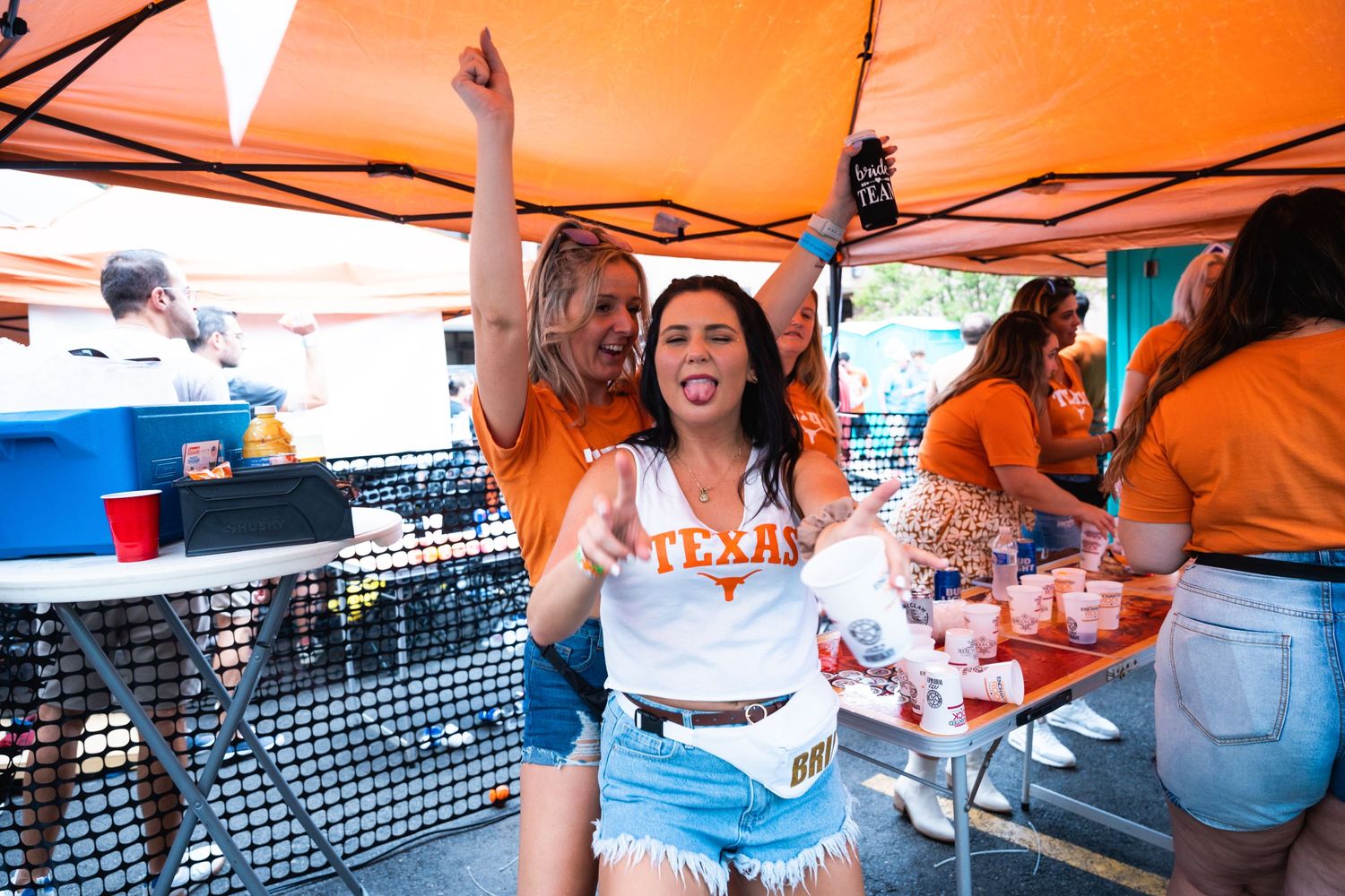 Excited party-goers celebrate with raised arms under orange tent at outdoor gathering.