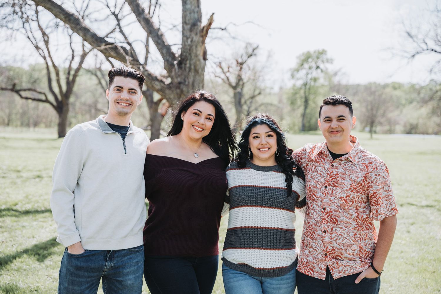 Group of four people smiling outdoors wearing casual attire with trees in background.
