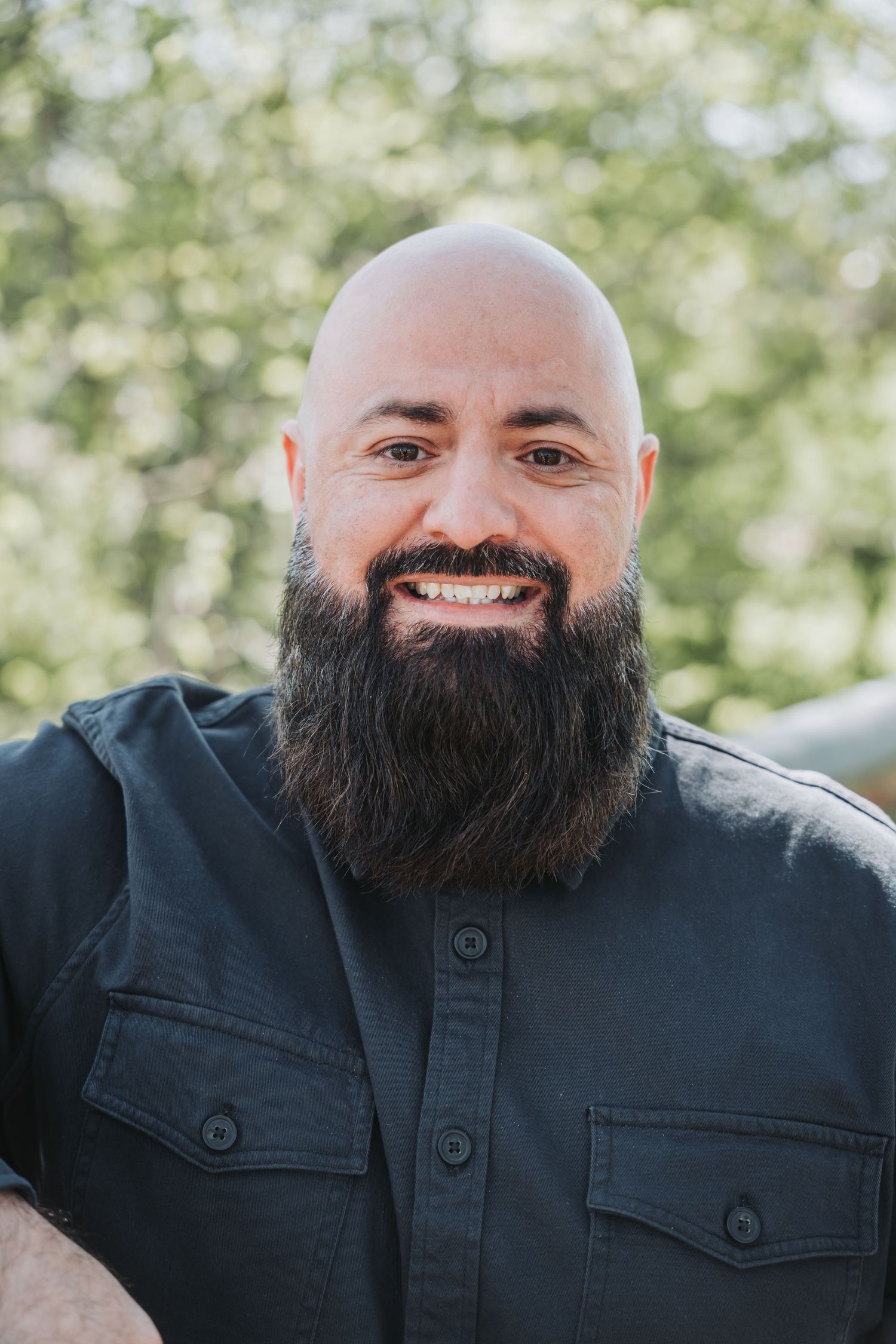 Portrait of a bald person with a full dark beard wearing a black button-up shirt outdoors.