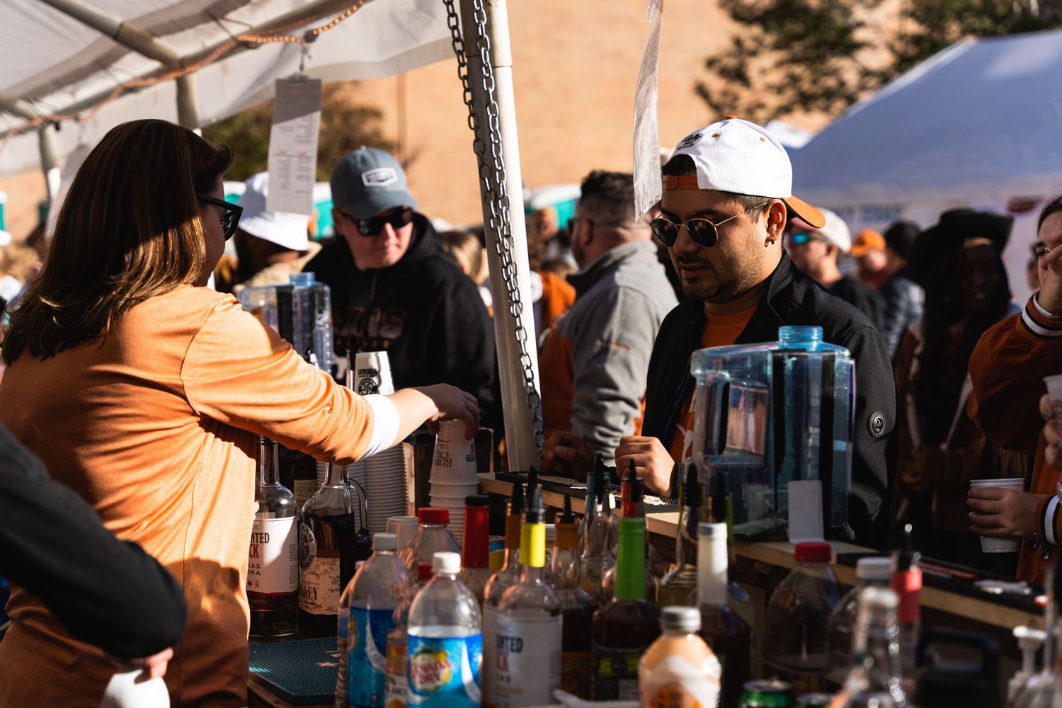 Bartenders serving drinks at an outdoor event tent with various bottles and mixers on display.