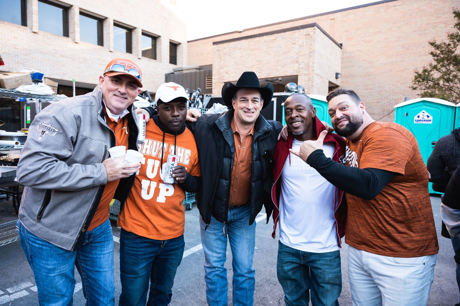 Sports fans in casual attire posing together while holding drinks at a tailgate party.