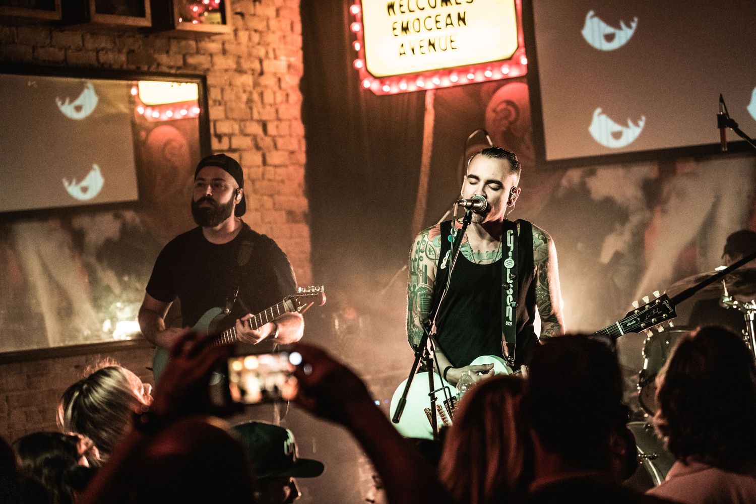 Rock band performing on stage with brick wall backdrop and illuminated Twitter bird decorations.