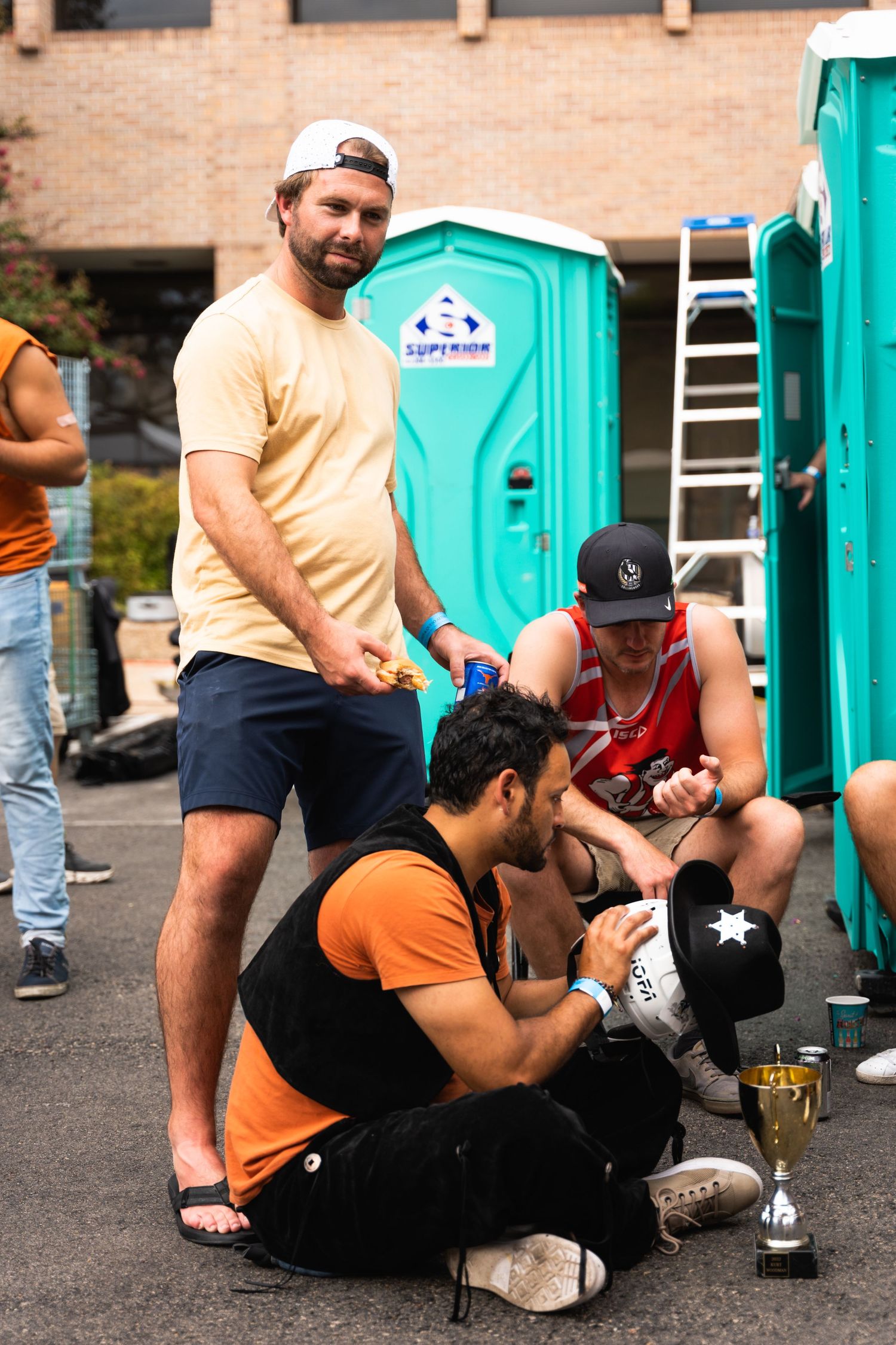 Group relaxing near portable toilets at outdoor festival with trophy nearby.