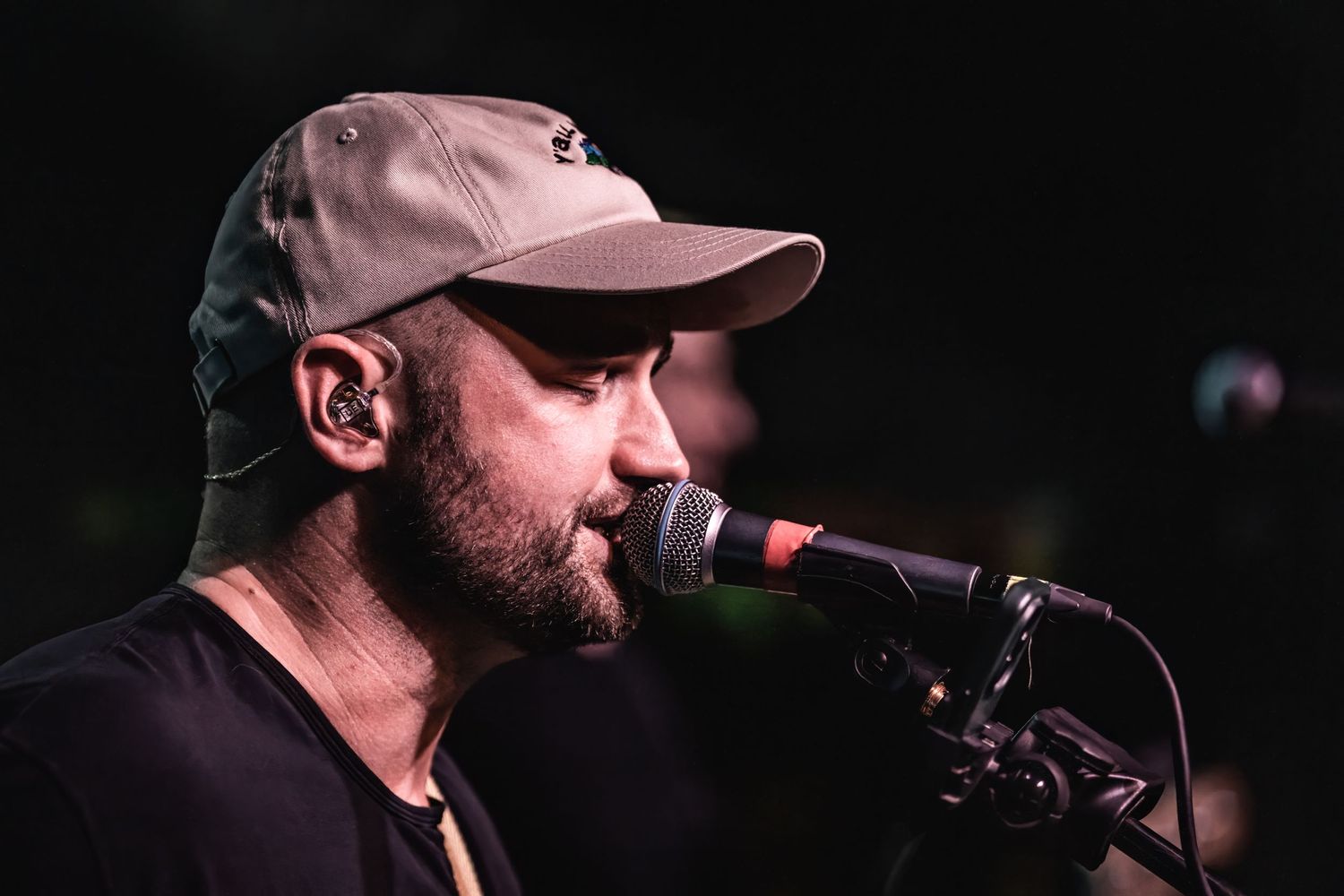 Singer in a baseball cap performs into microphone during intimate concert venue performance.