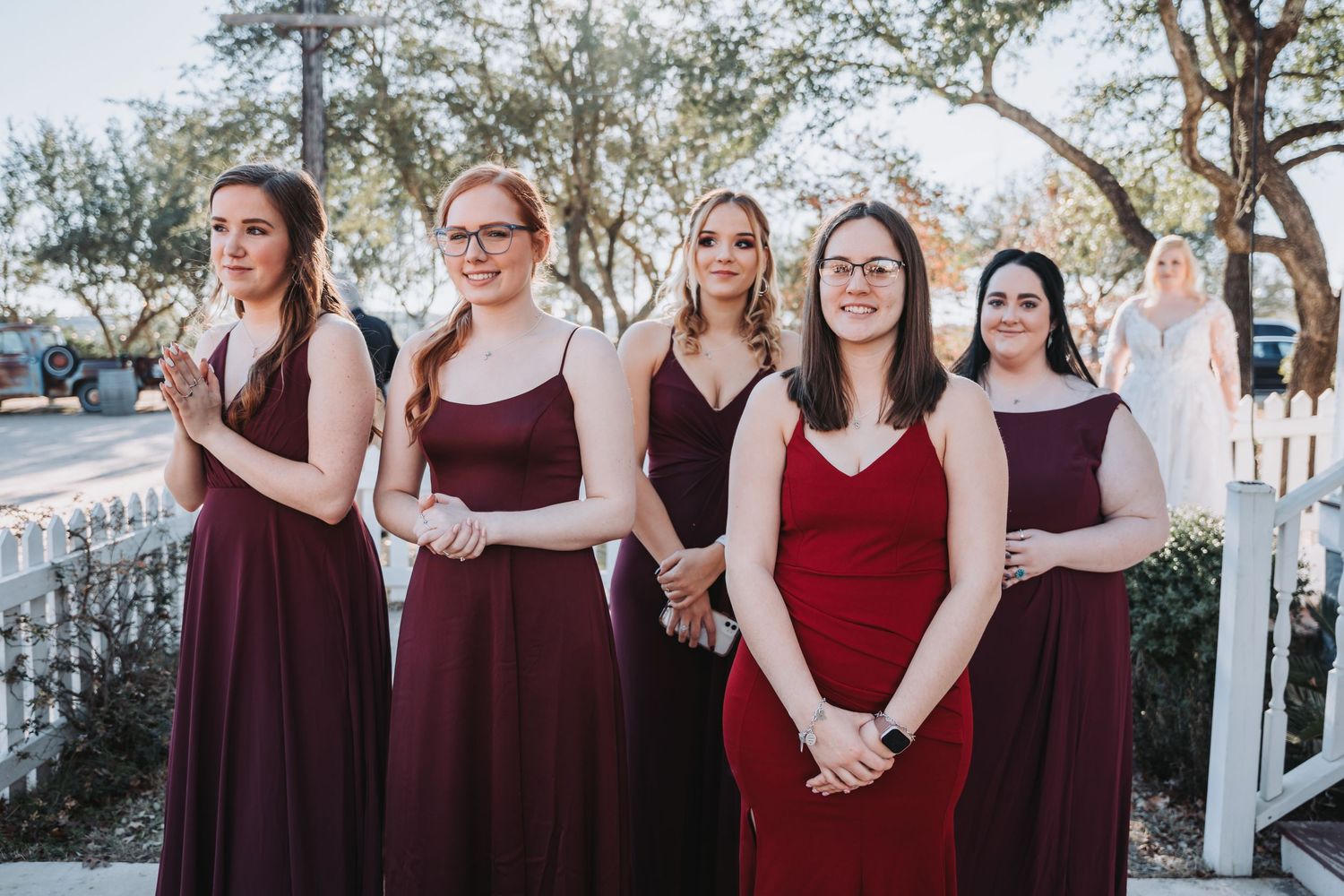 Bridesmaids in burgundy dresses stand together outdoors on a bright day.