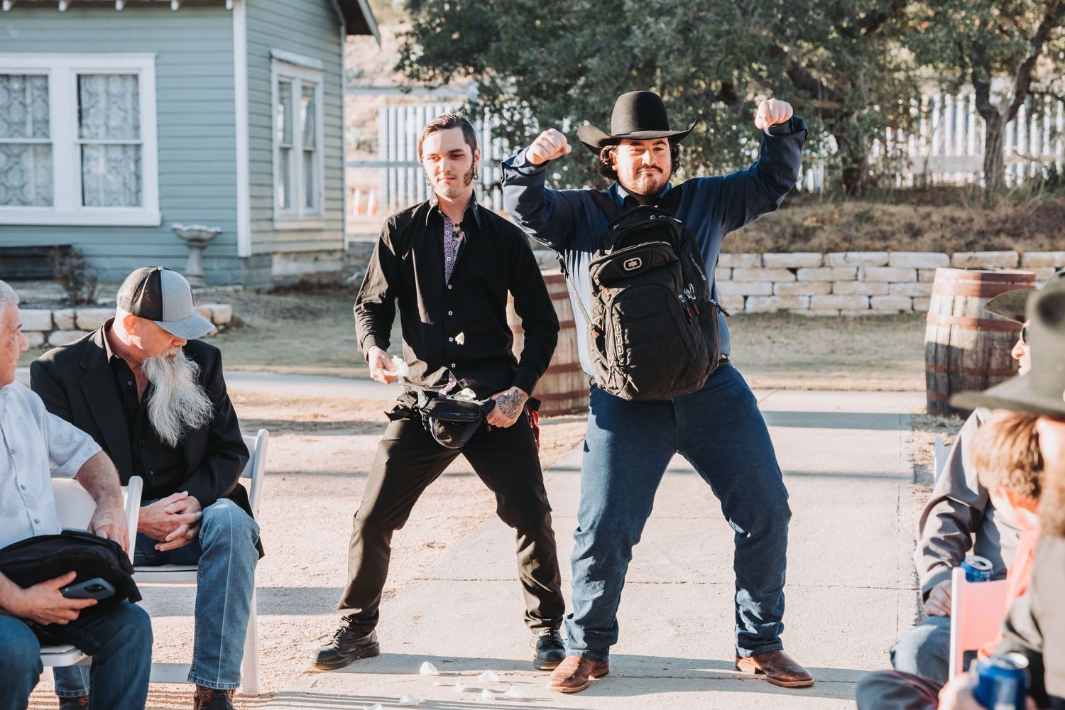 A group strikes playful poses during outdoor wedding festivities with rustic buildings in background.