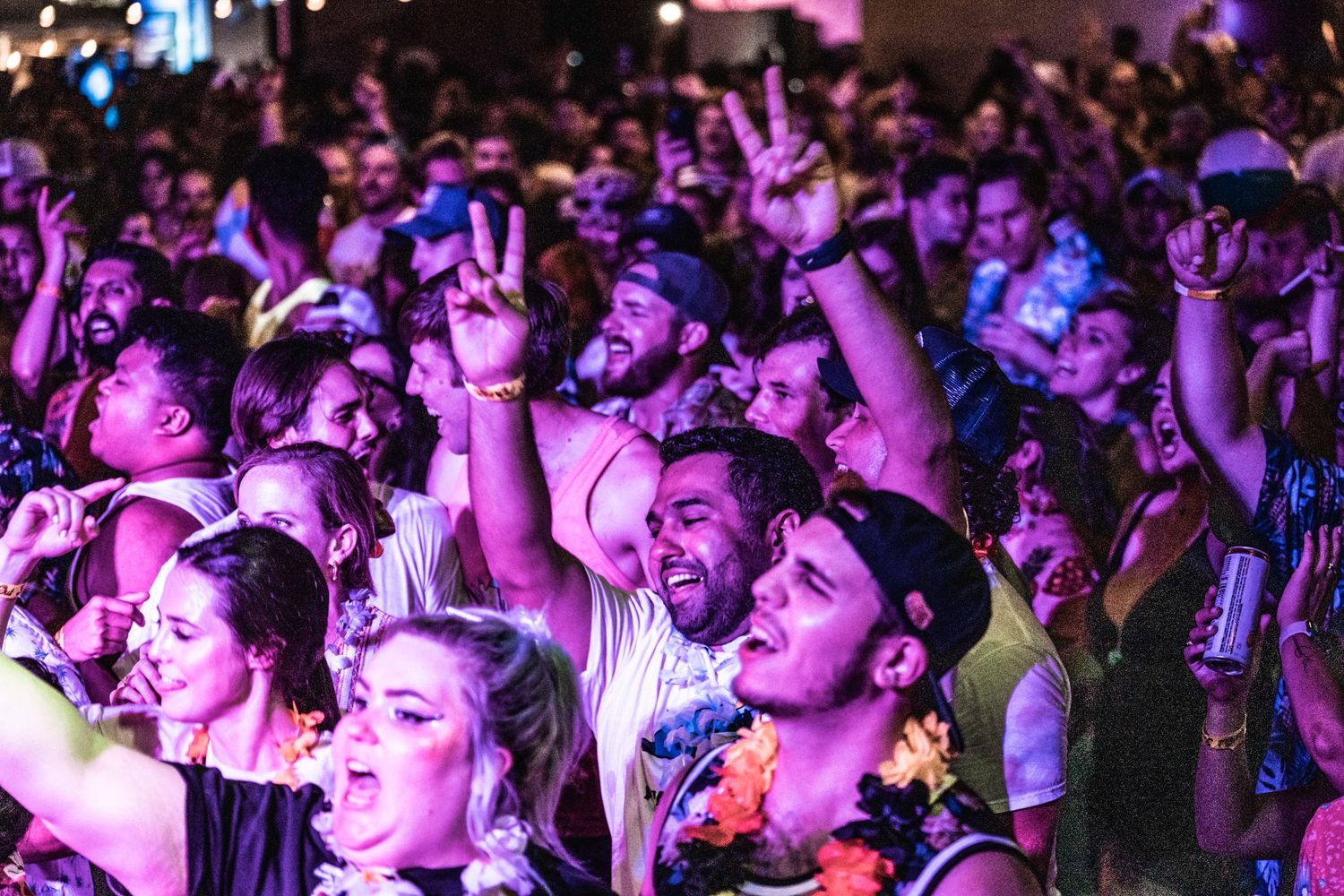 A crowded concert audience cheering and raising hands under purple lighting.