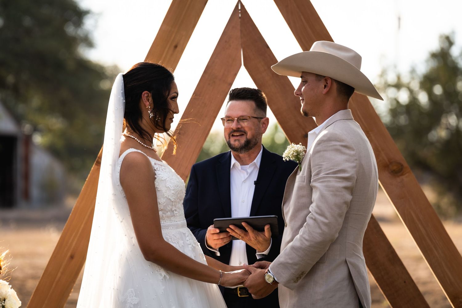 A wedding ceremony takes place under a wooden triangular arch at sunset.
