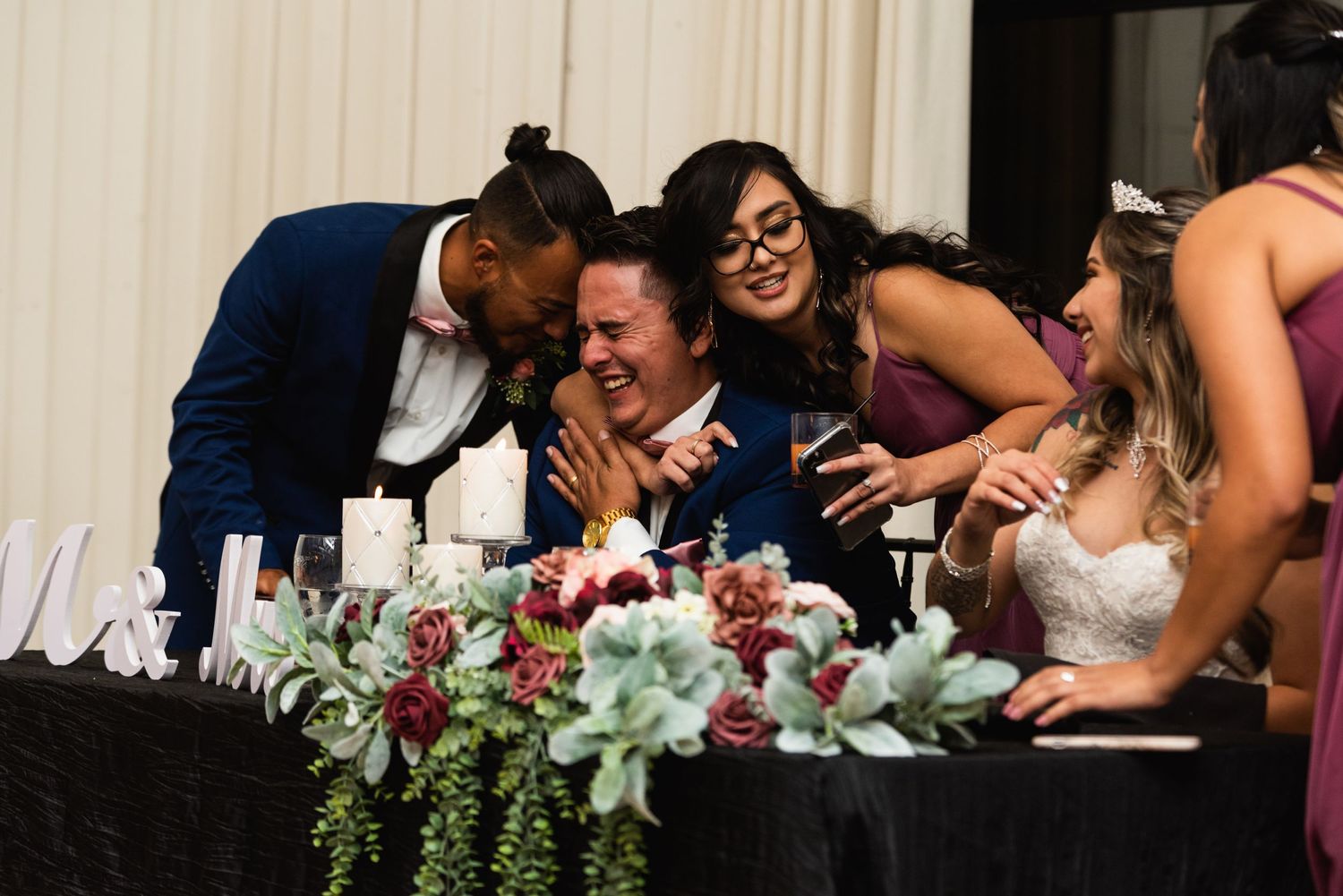 Wedding party sharing a candid laughing moment at the reception head table with floral decorations.