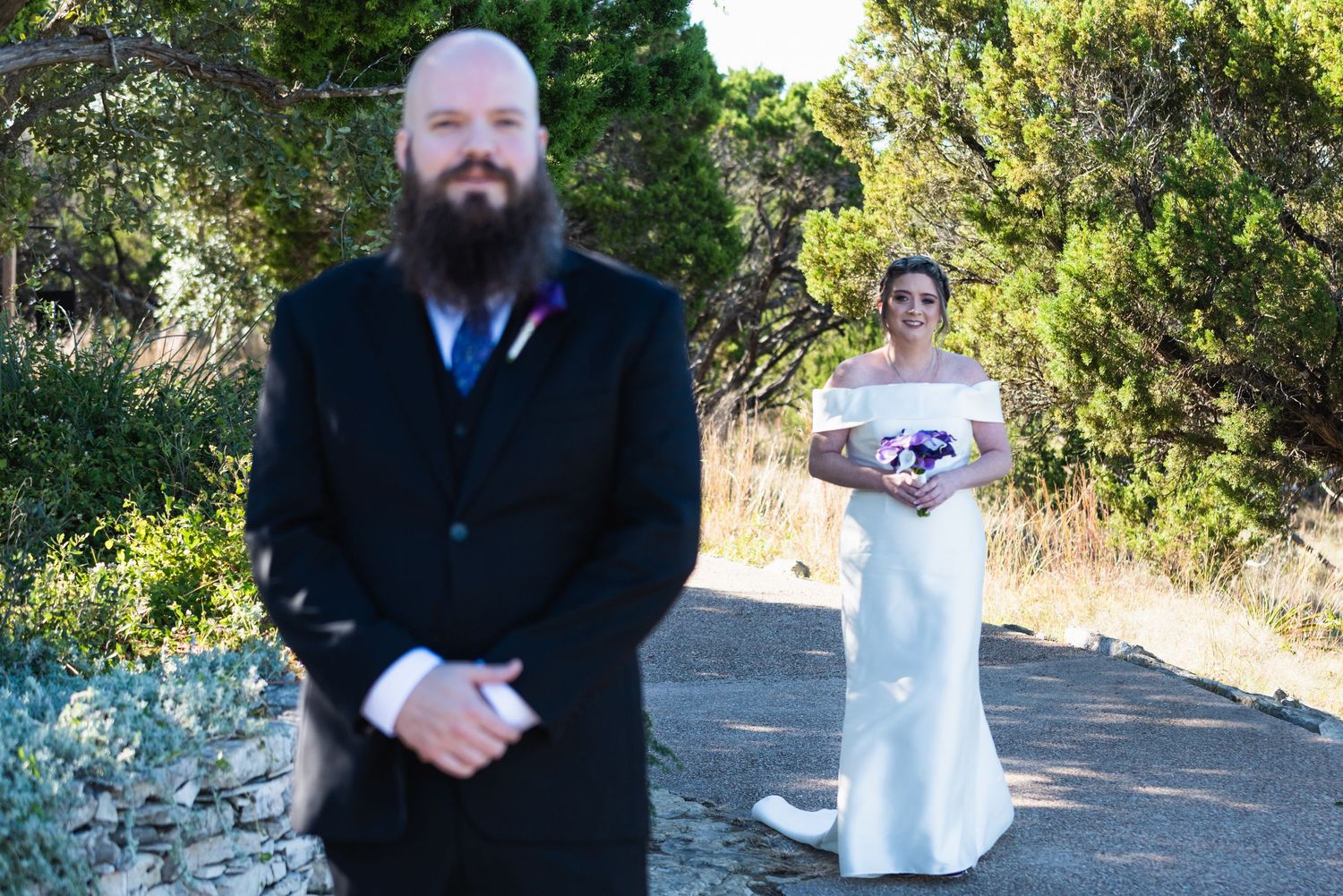 First look wedding photo with groom in foreground and bride standing in background on path.