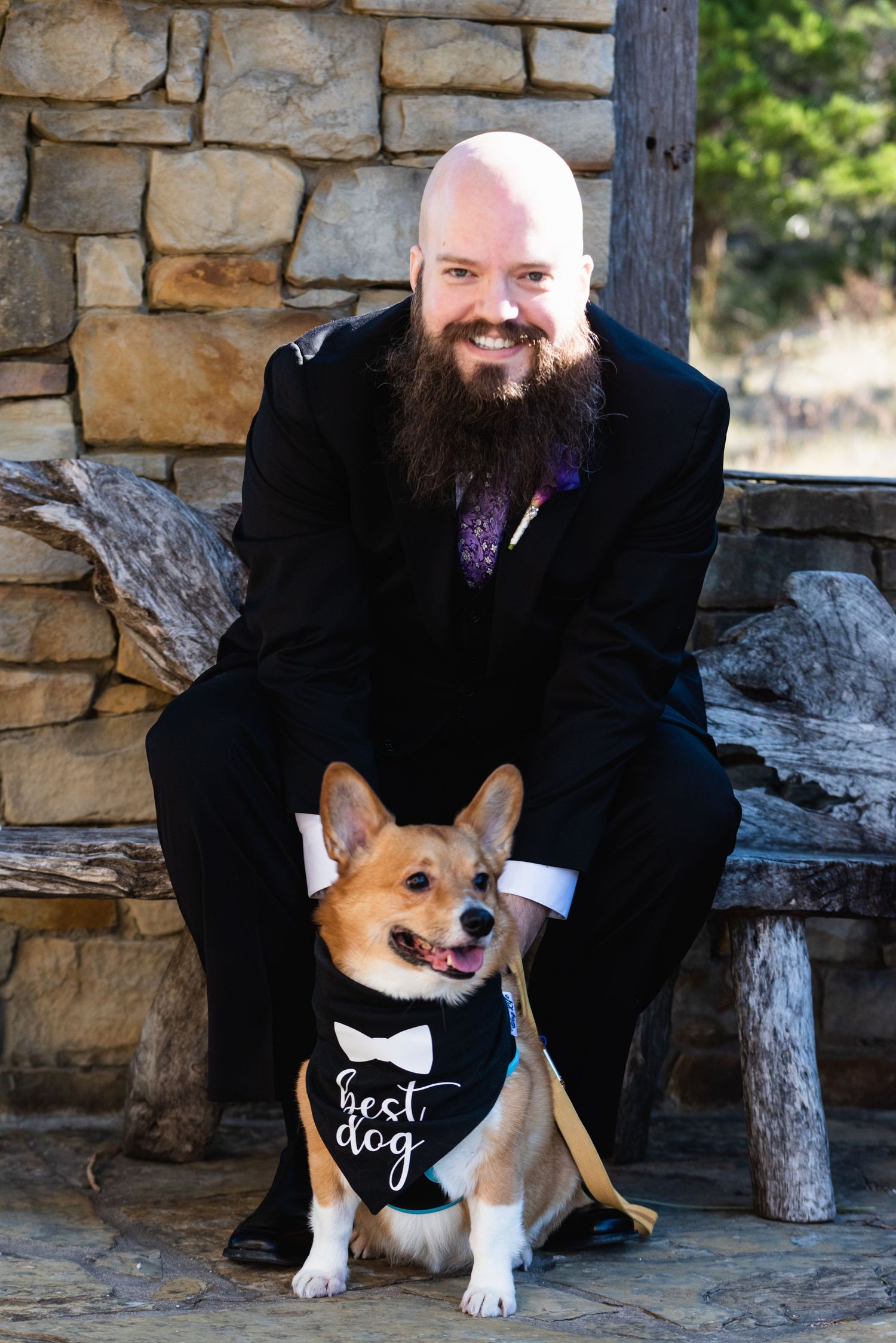 A bearded man in a suit poses with a corgi wearing a 'First Dog' bowtie bandana.