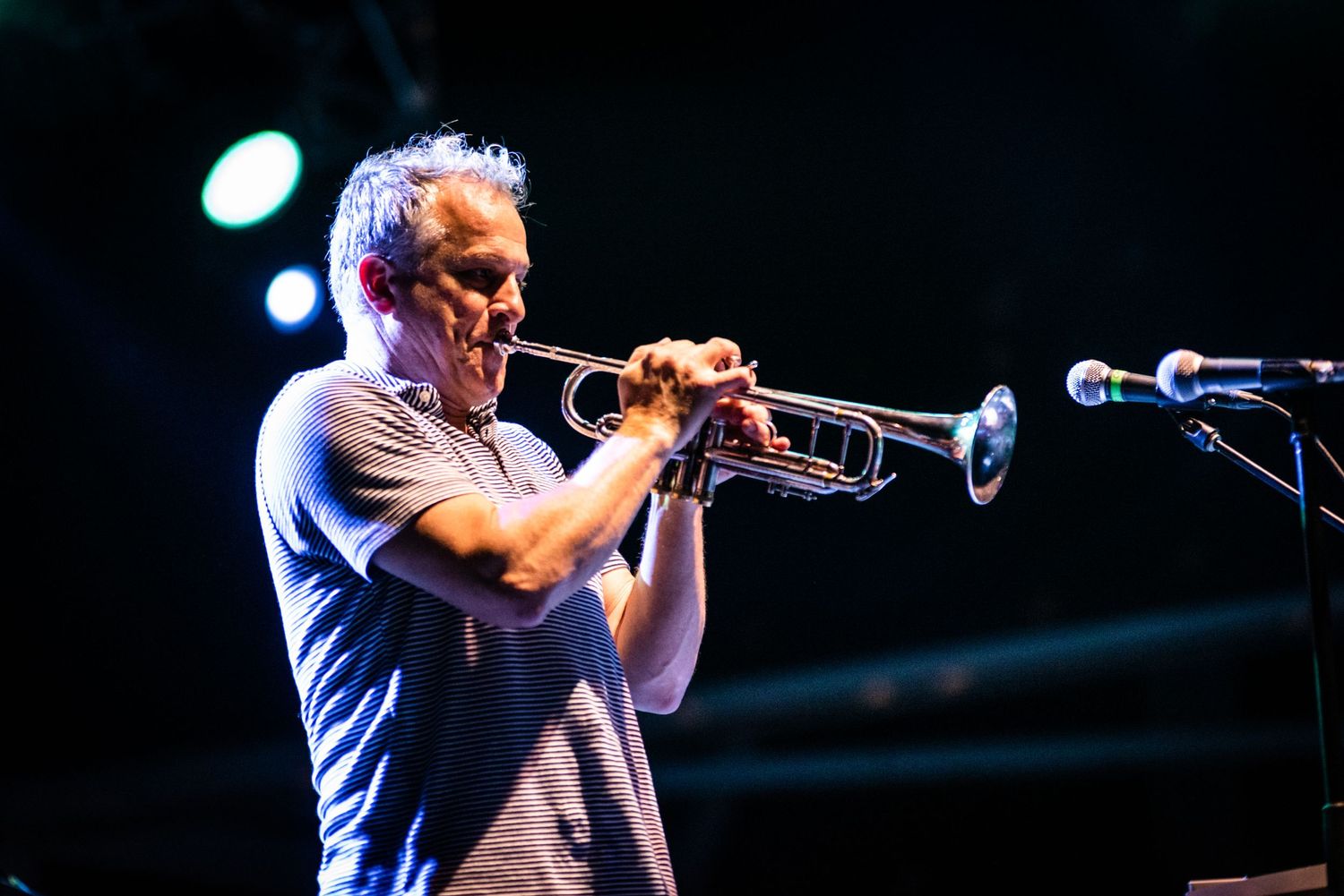 Musician playing trumpet on stage with dramatic blue lighting and microphone.
