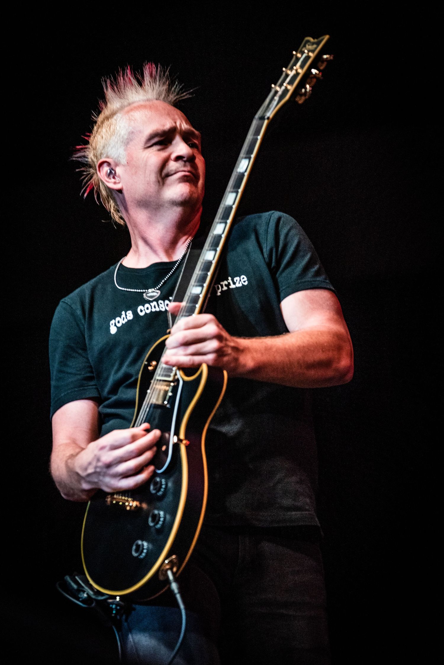 Rock guitarist with spiky hair performs with black Les Paul guitar on dark stage.