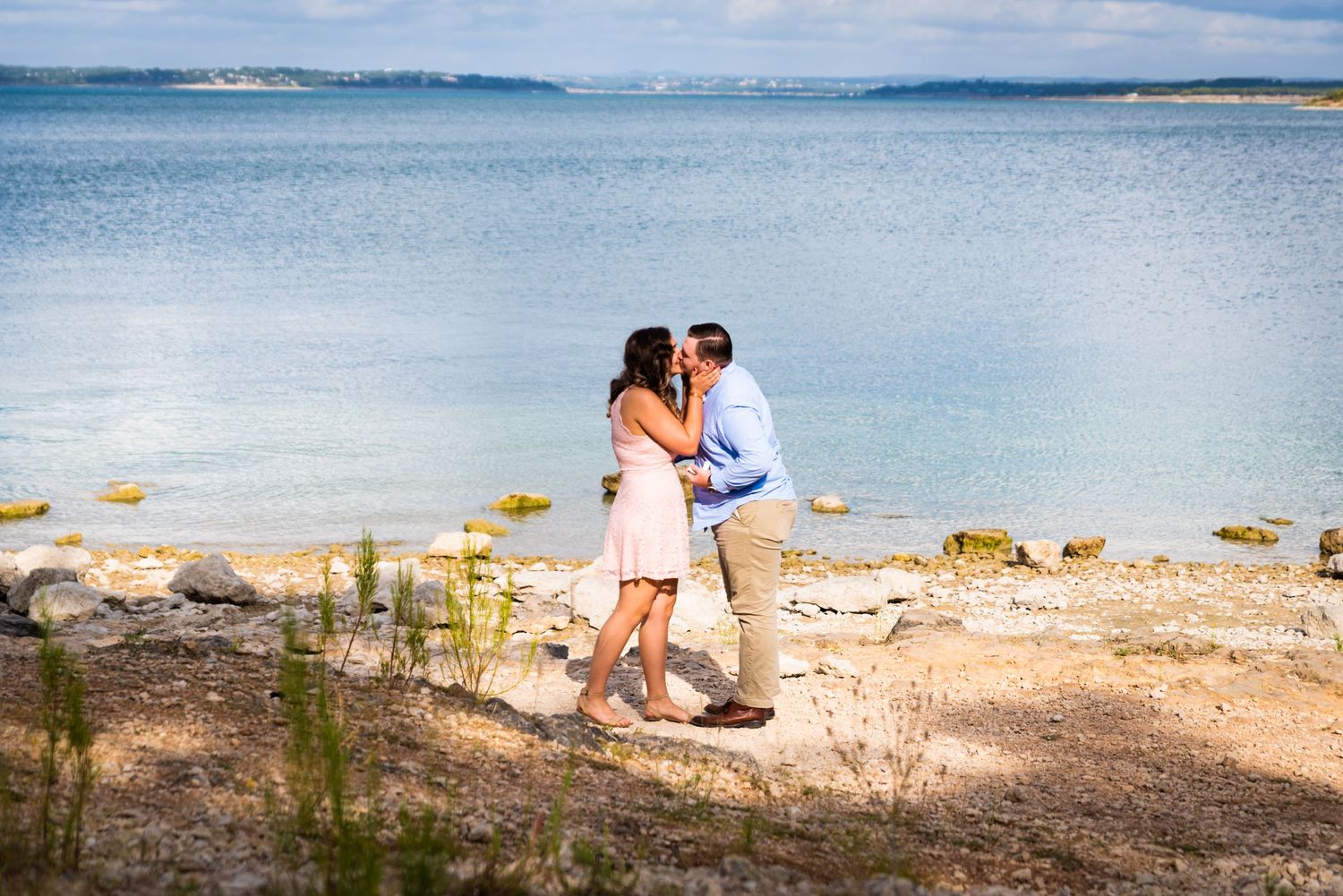 A romantic couple shares a tender moment on a rocky beach overlooking calm blue waters.