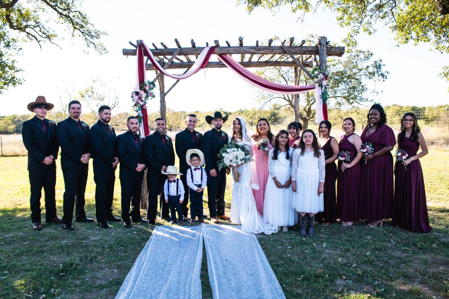 Wedding party stands under wooden pergola decorated with red fabric in an outdoor rustic ceremony setting.