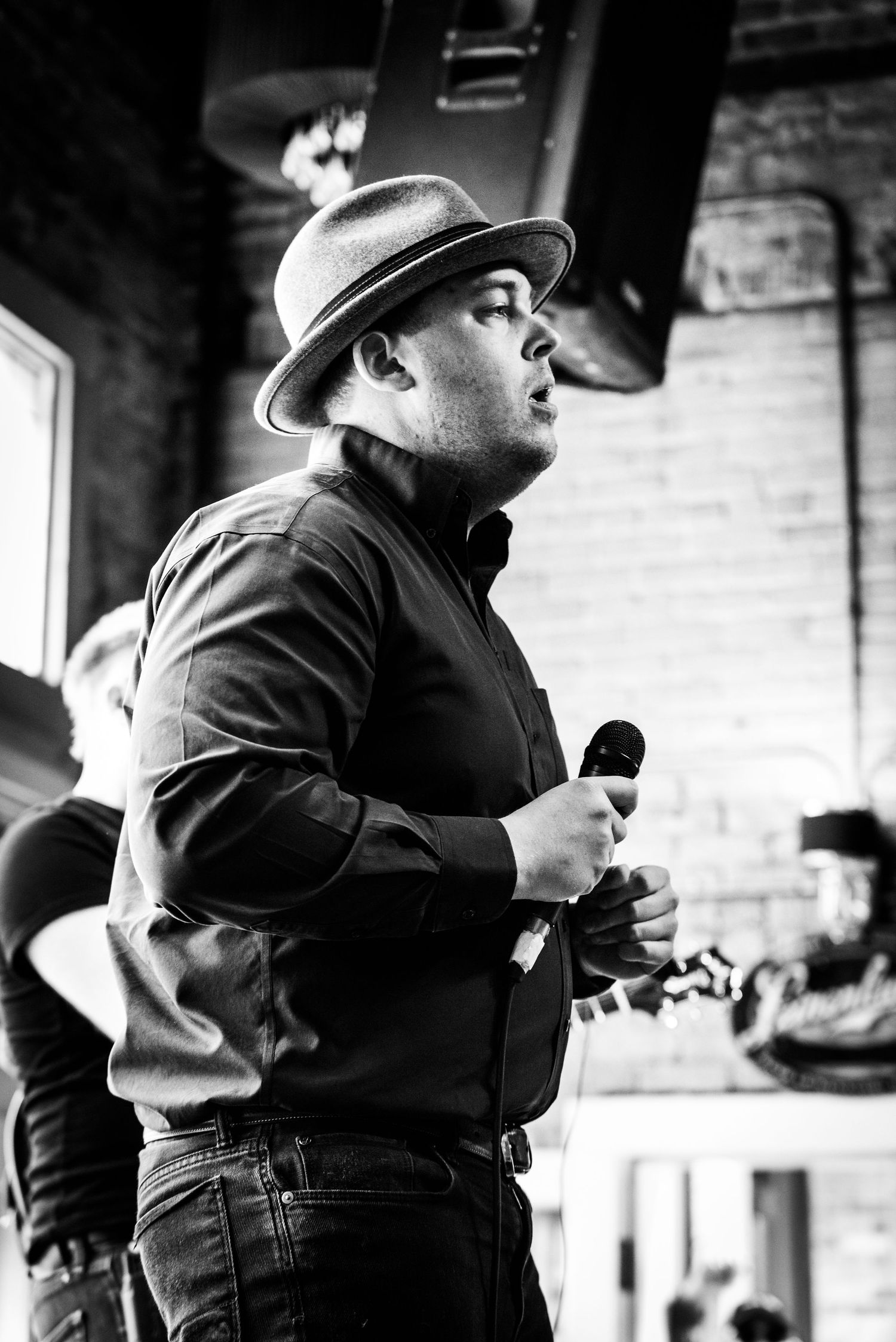 A performer in a fedora singing into a microphone against a brick wall background.