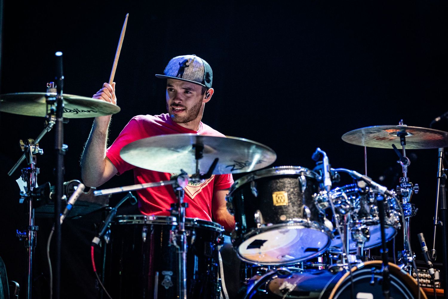 Drummer with baseball cap plays drum kit during live performance with colored stage lighting.
