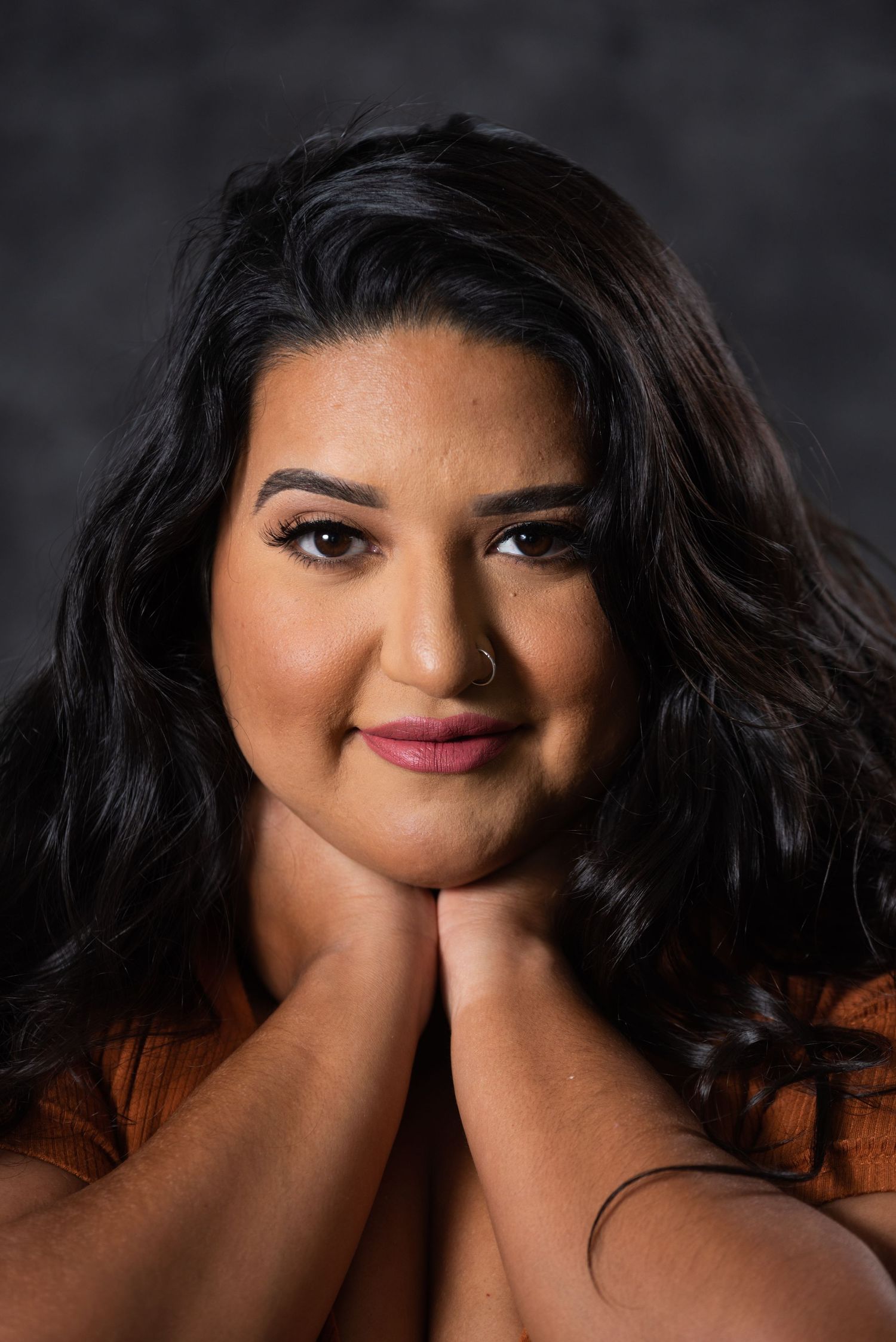 Studio portrait of woman with flowing dark hair wearing orange top against dark backdrop.