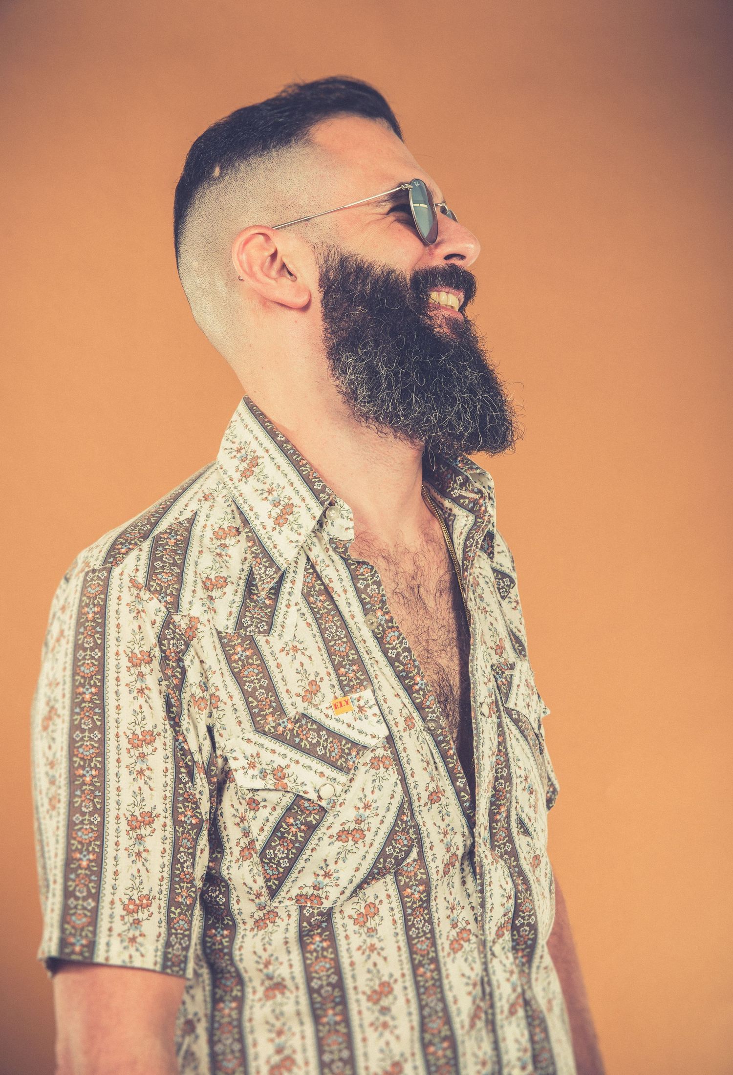 Studio portrait of a bearded person in a patterned short sleeve button-up shirt against an orange background.