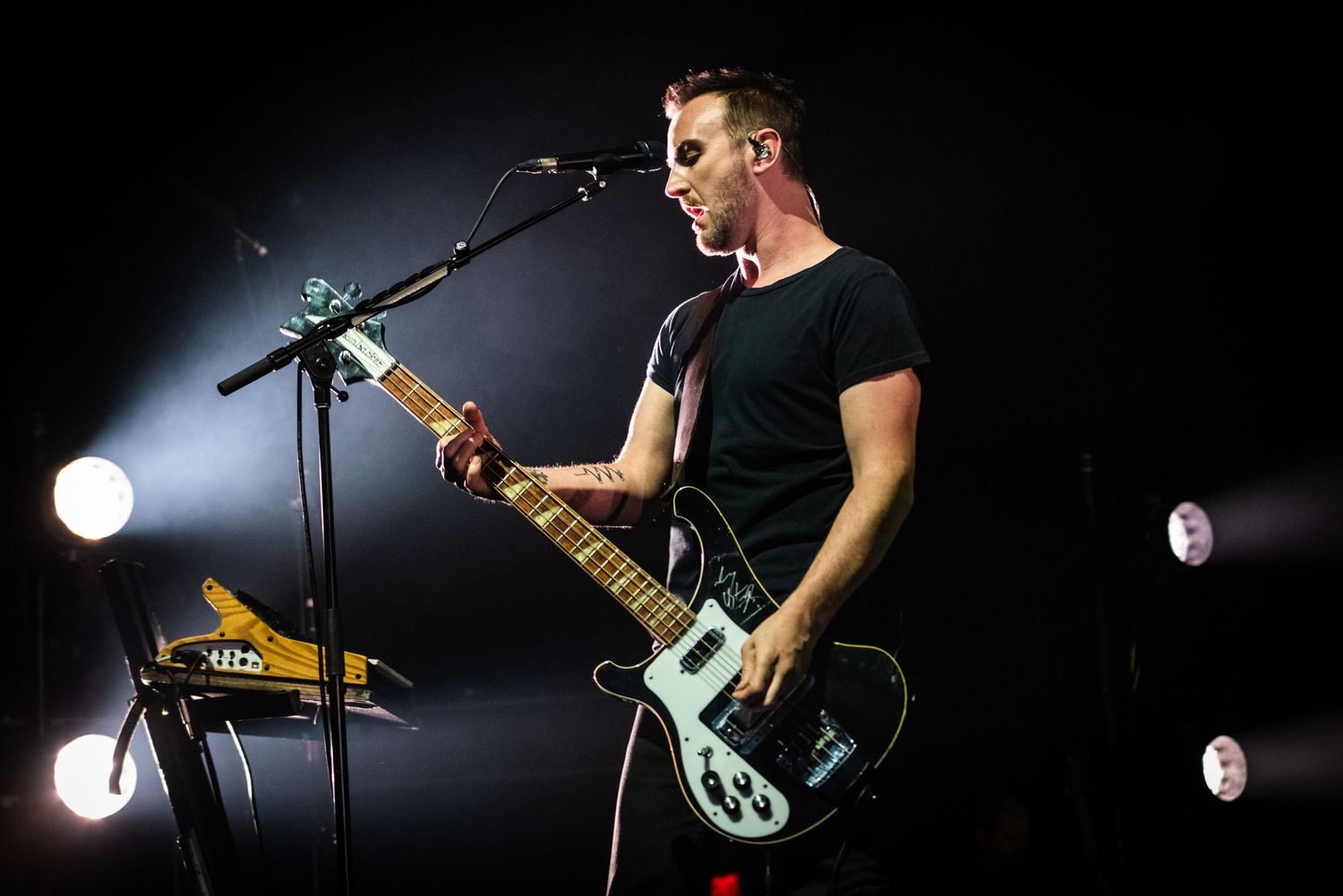 Guitarist plays electric guitar while singing into microphone on darkened stage with spotlights.