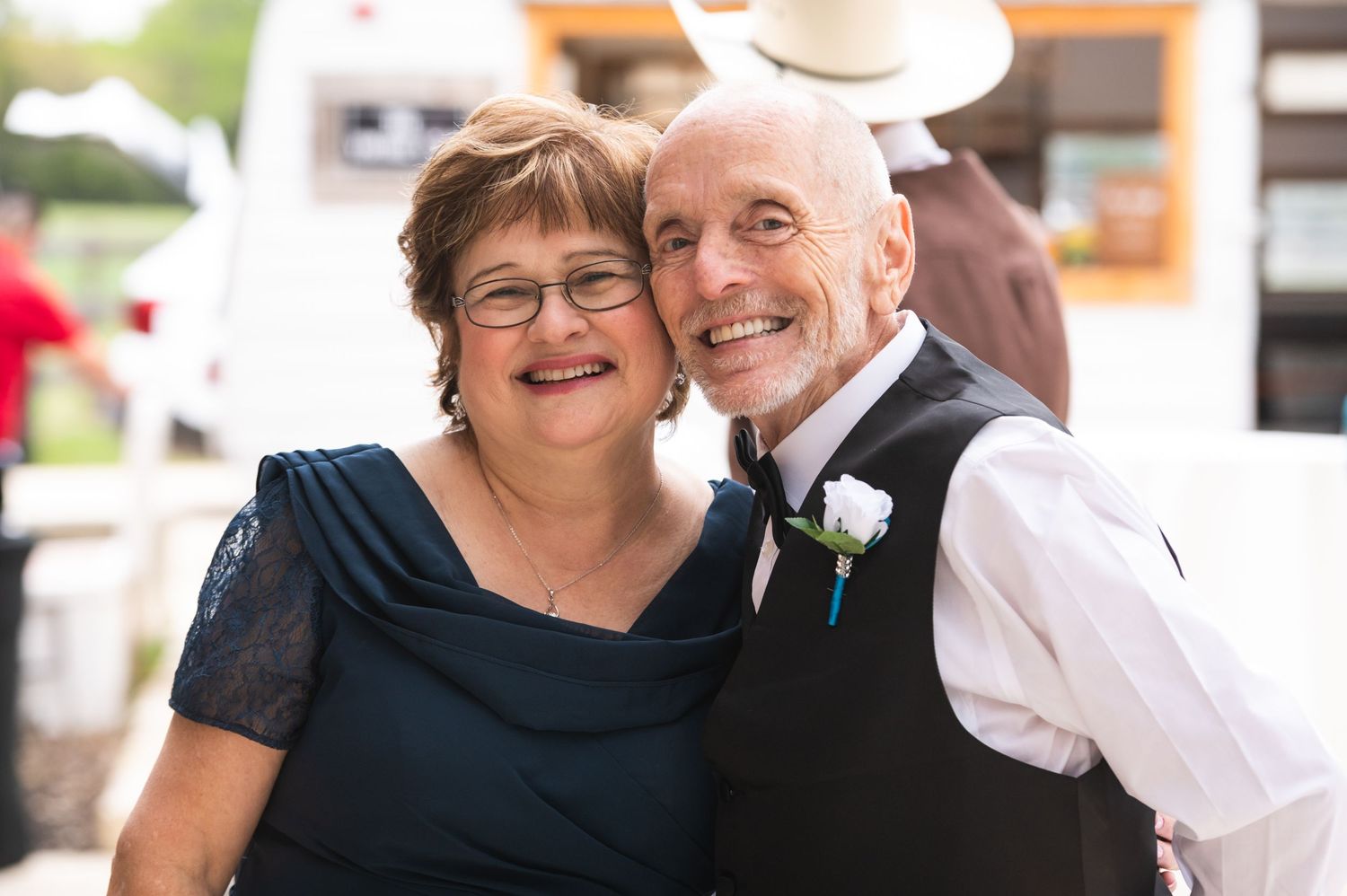 A mature couple in formal attire share a joyful moment at a wedding celebration, with the groom wearing a vest and boutonniere.