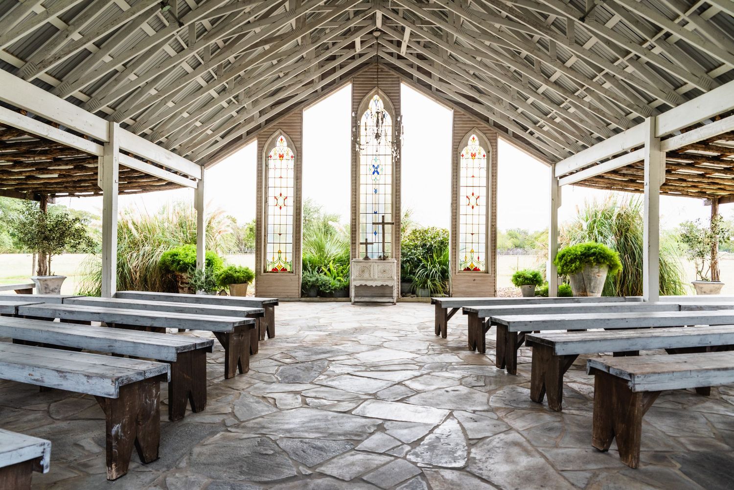 Open-air chapel with wooden benches and stained glass windows in a rustic outdoor setting.