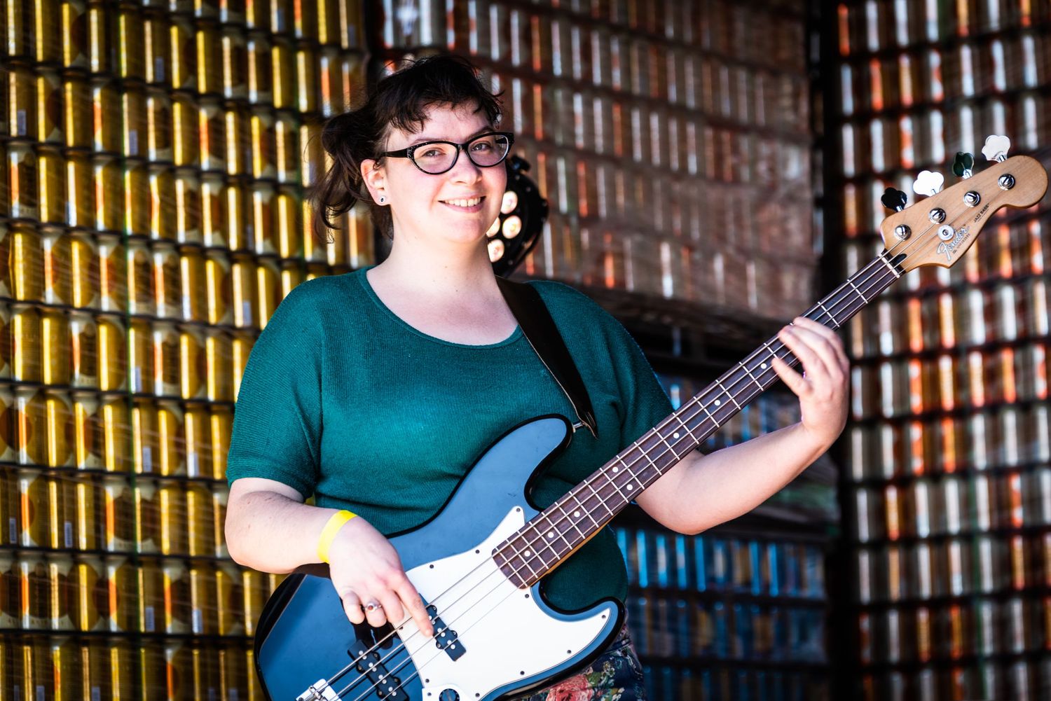 Bass player in green shirt performs against illuminated bottle wall backdrop.