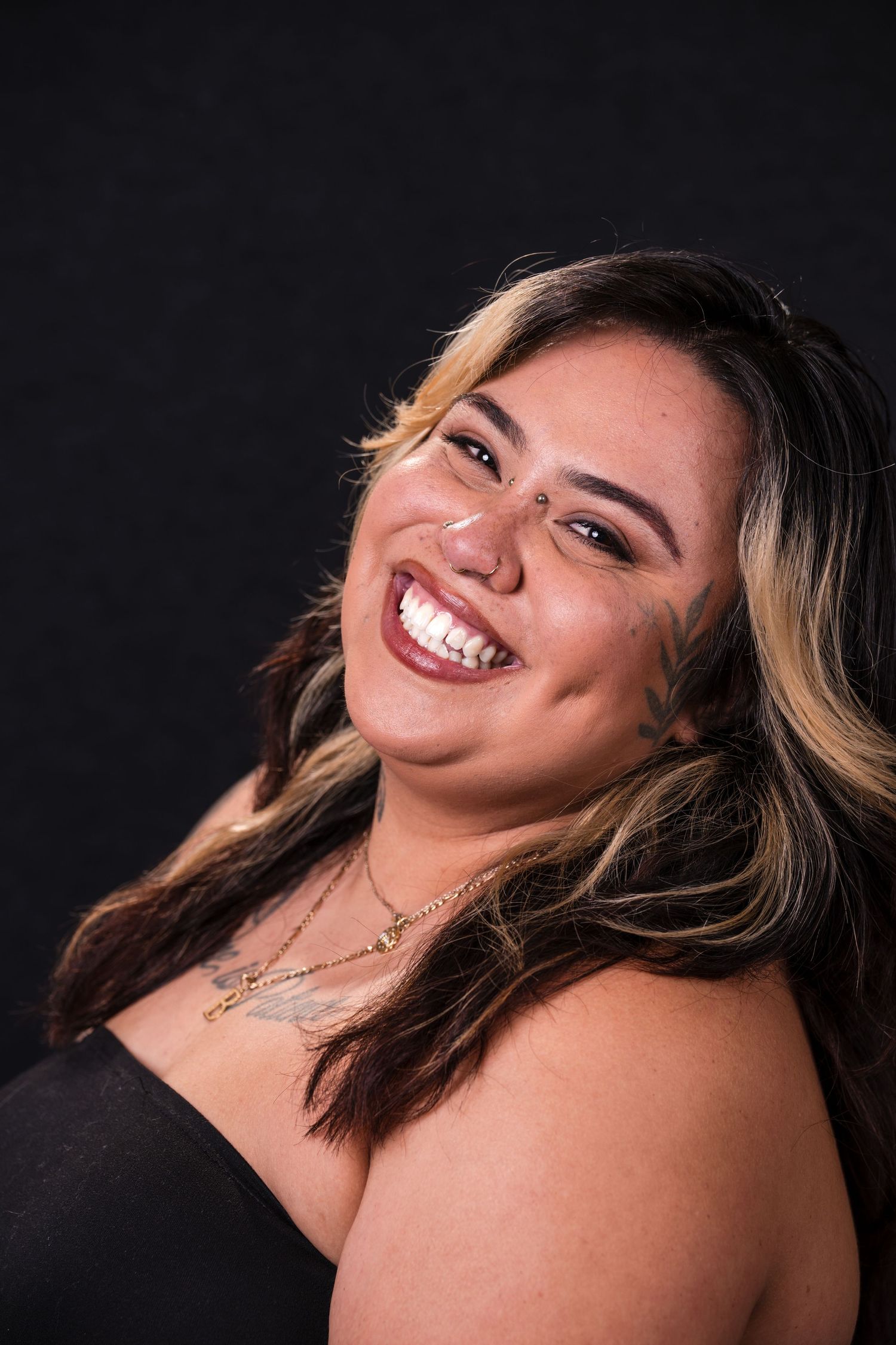 Portrait of someone with blonde highlights wearing black against dark background, showing bright genuine smile.