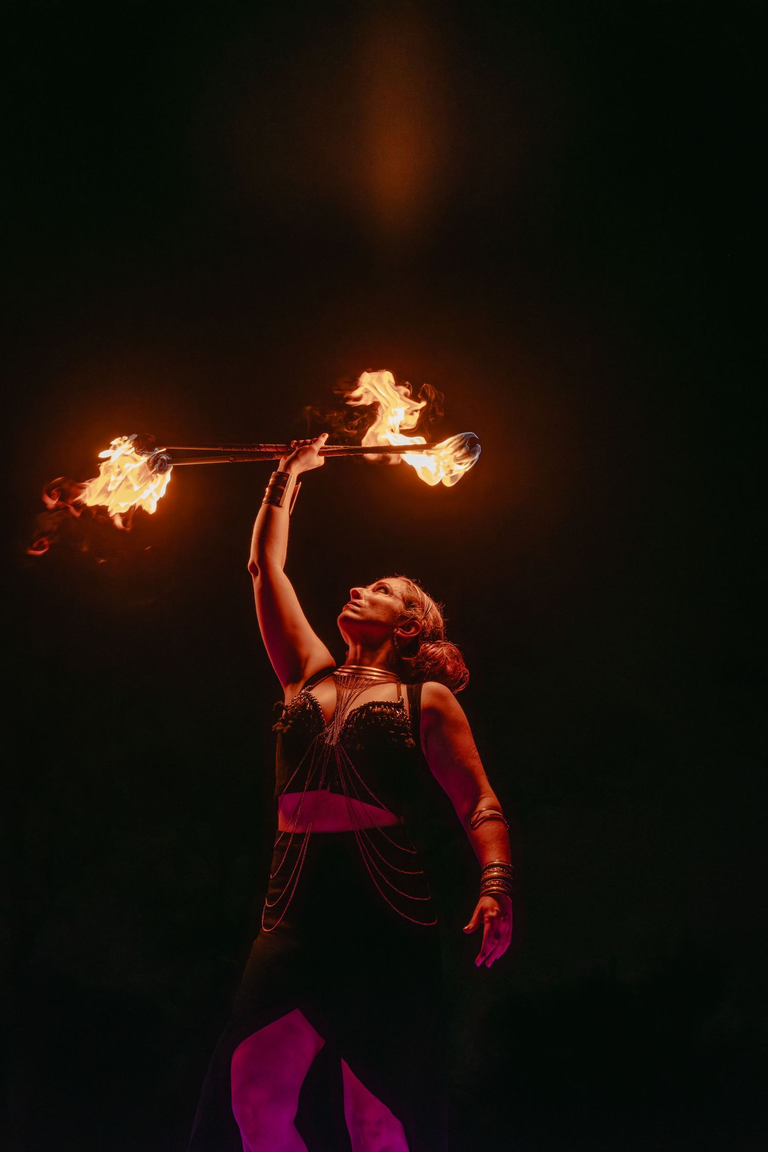 A fire dancer performs with a flaming staff against a dark background, flames illuminating her dramatic pose.