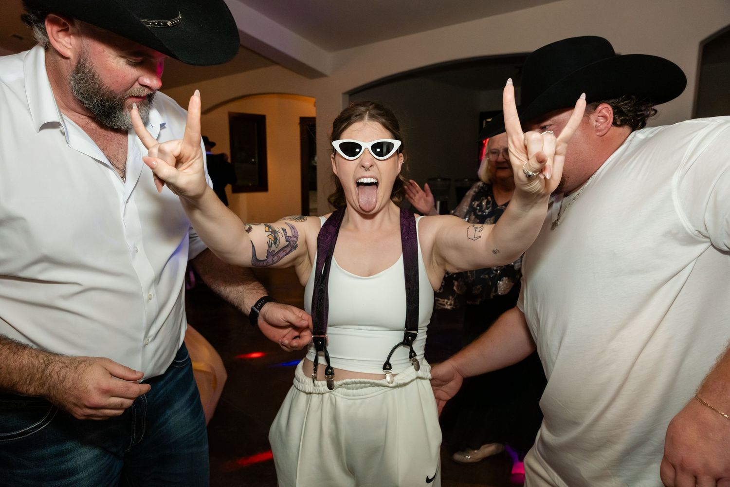 Three people dancing at a party, woman in white sunglasses and suspenders making rock signs with two men in black hats.