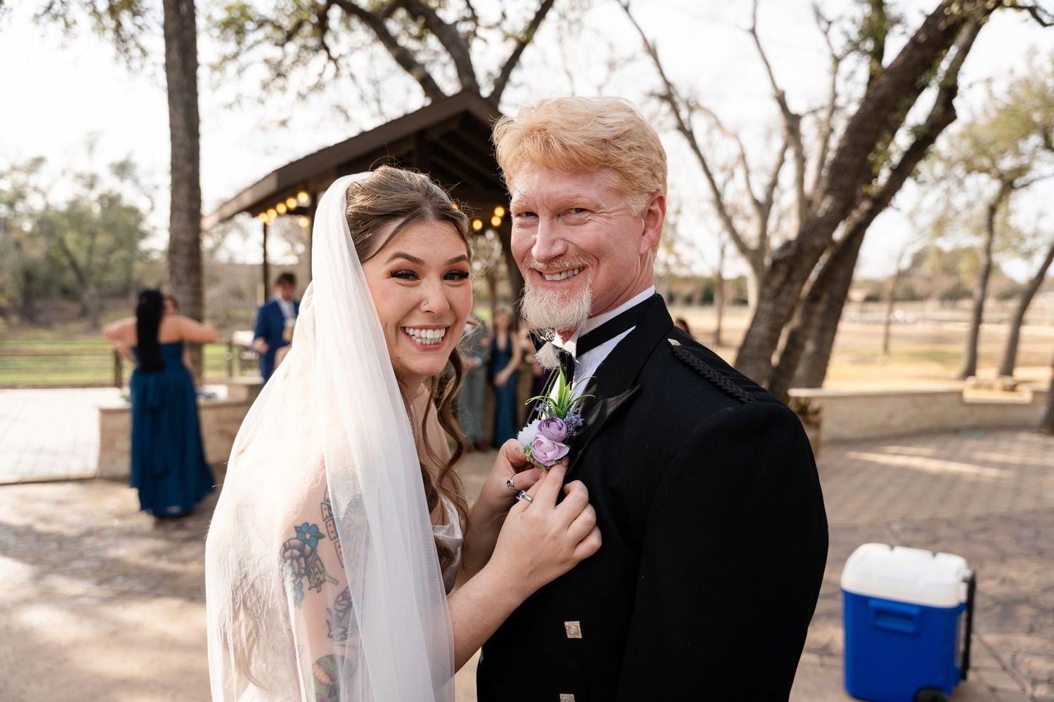 Smiling bride in white veil adjusts groom's boutonniere outdoors at rustic wedding venue with trees in background.