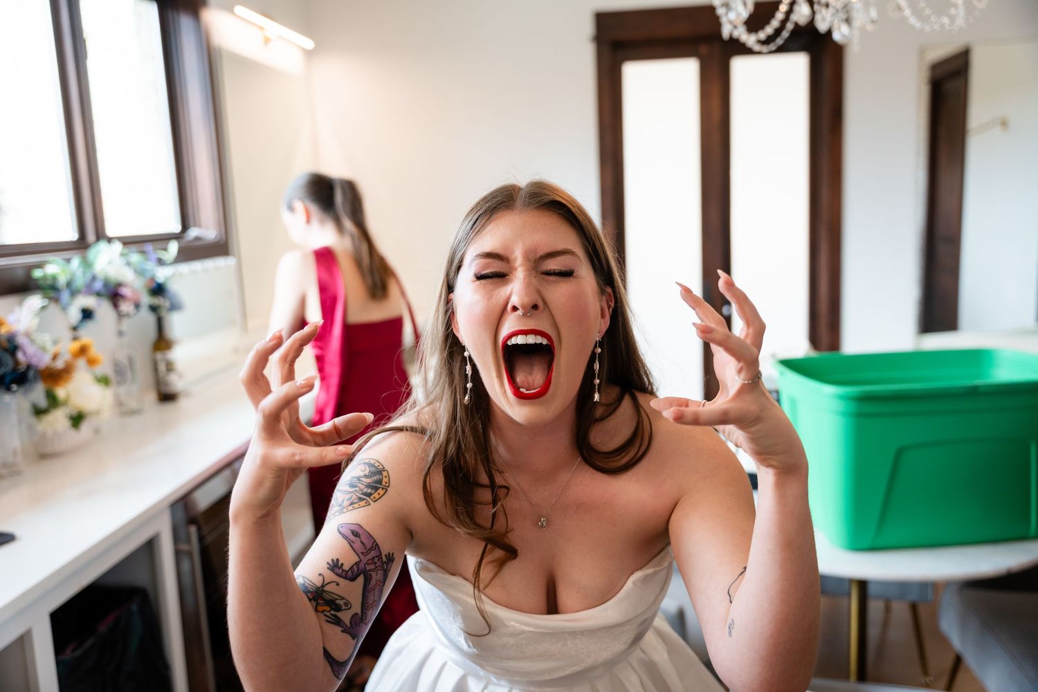 Bride in white dress screaming excitedly with hands raised, tattoos visible, woman in red dress in background.