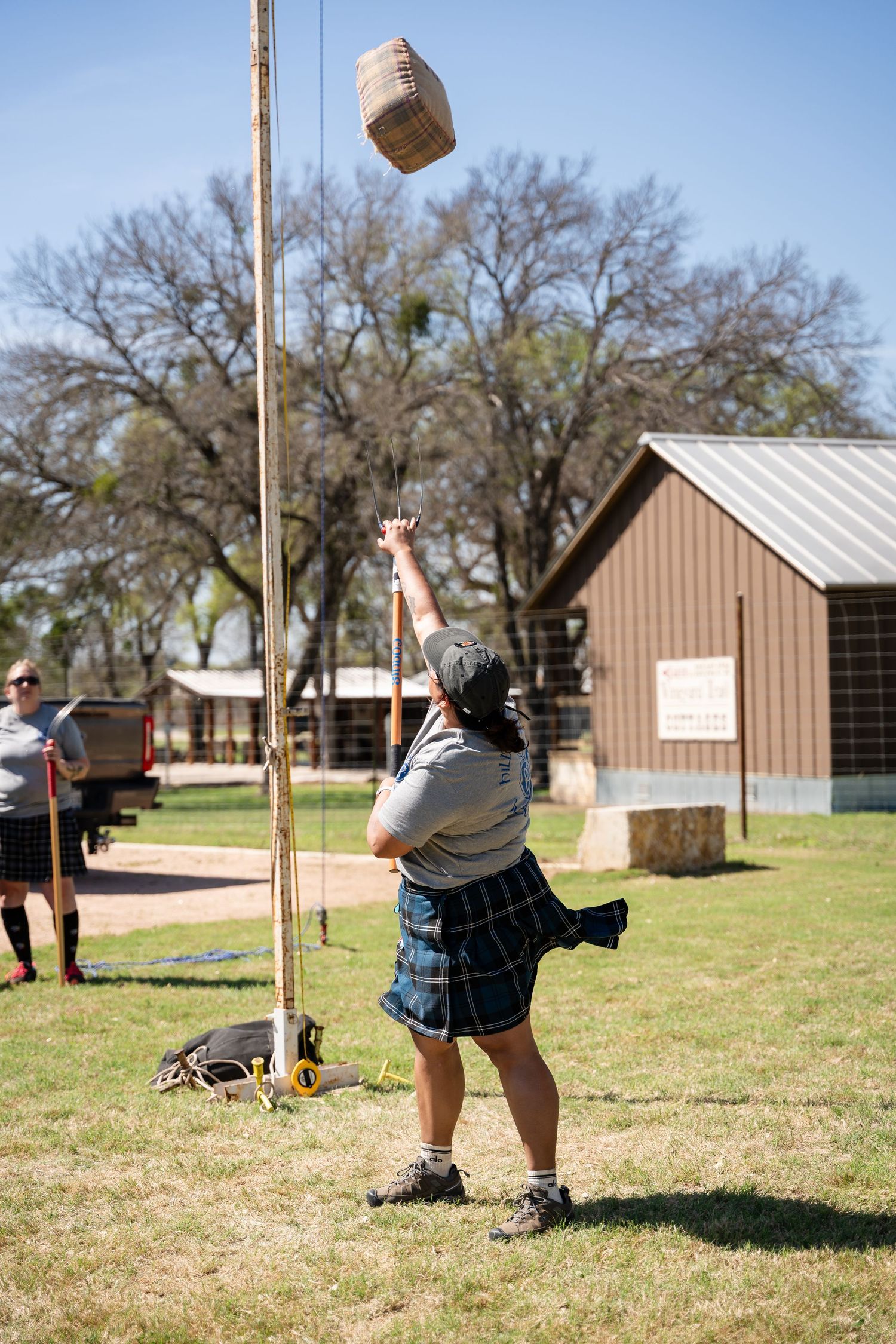 Athlete in kilt throws weighted bag over bar at Highland Games competition outdoors.