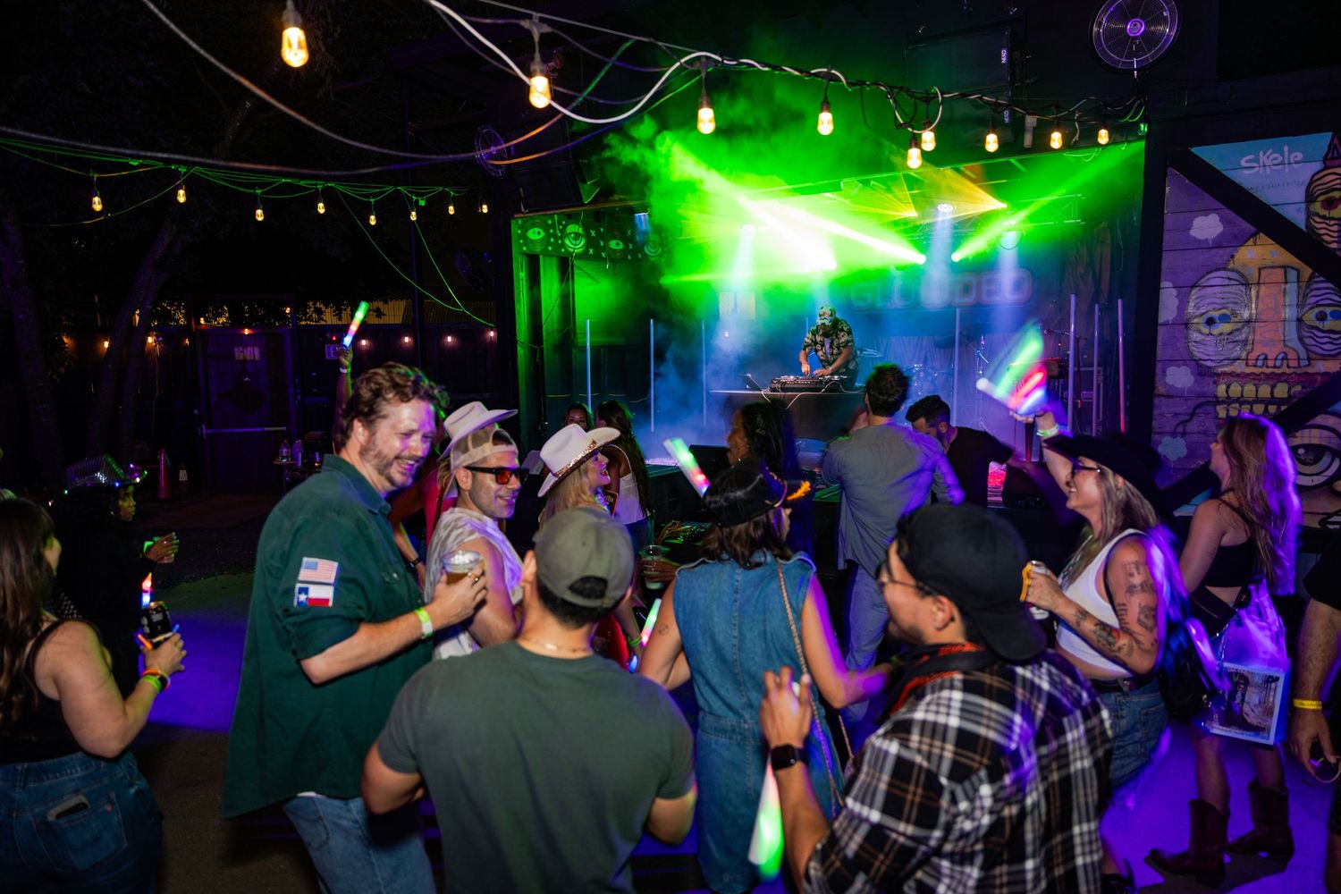 Crowd of people dancing at a lively nightclub with green stage lights and a DJ performing in the background.
