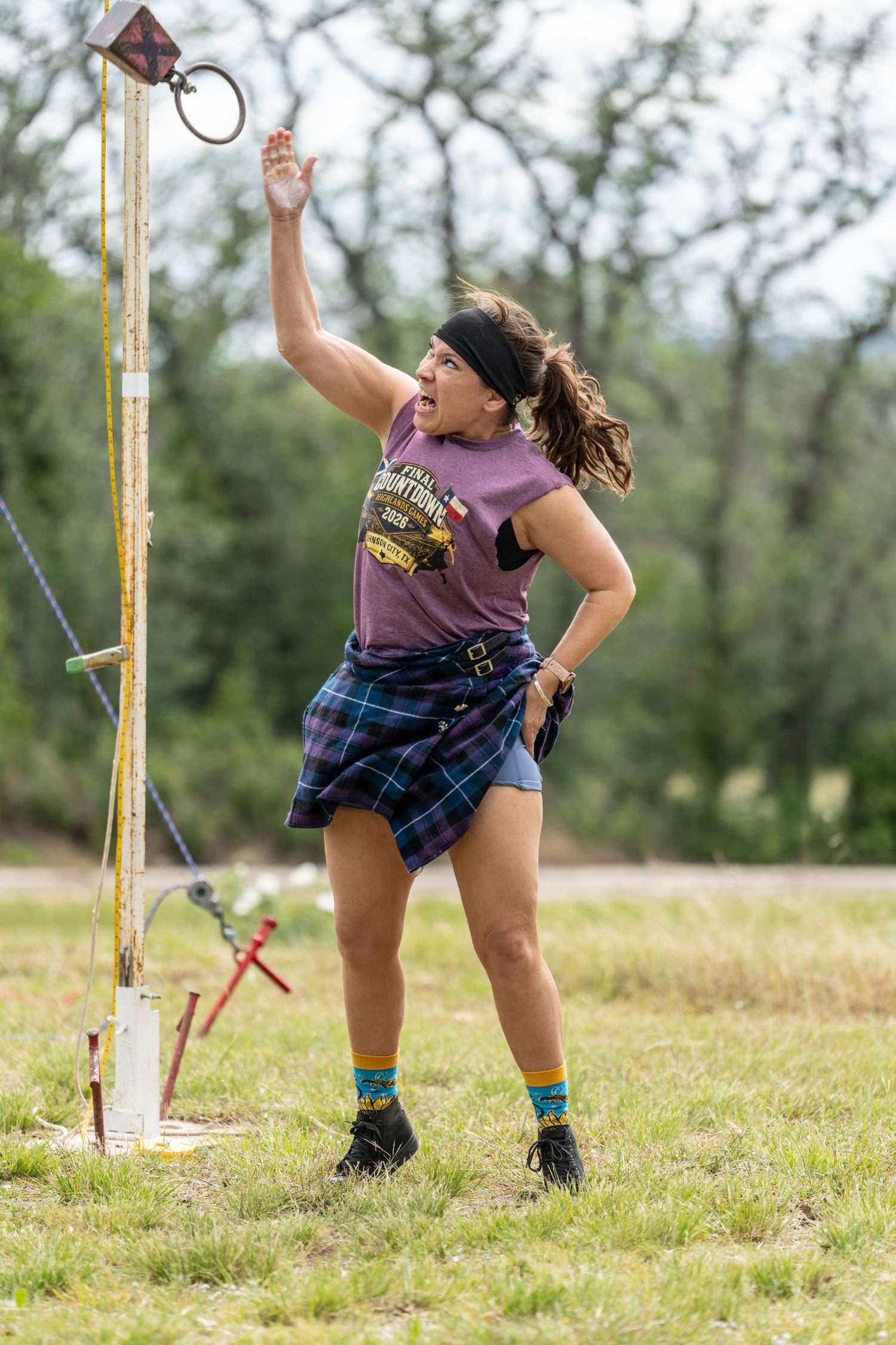 Woman in tartan kilt and purple shirt competing in Highland Games, reaching up toward a pole on grassy field.