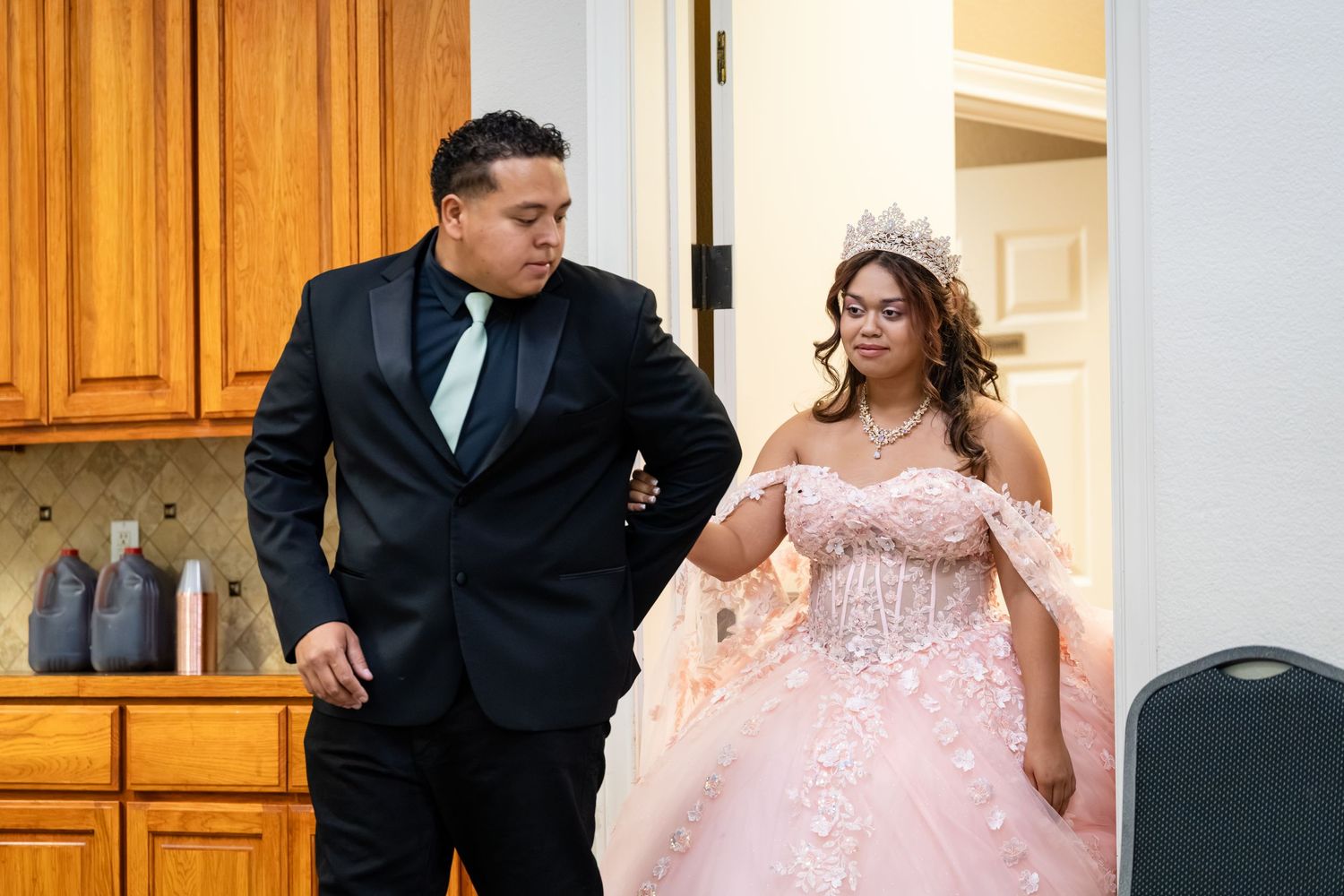 A young couple in formal attire, man in black suit and girl in pink quinceañera gown with tiara, entering a celebration.