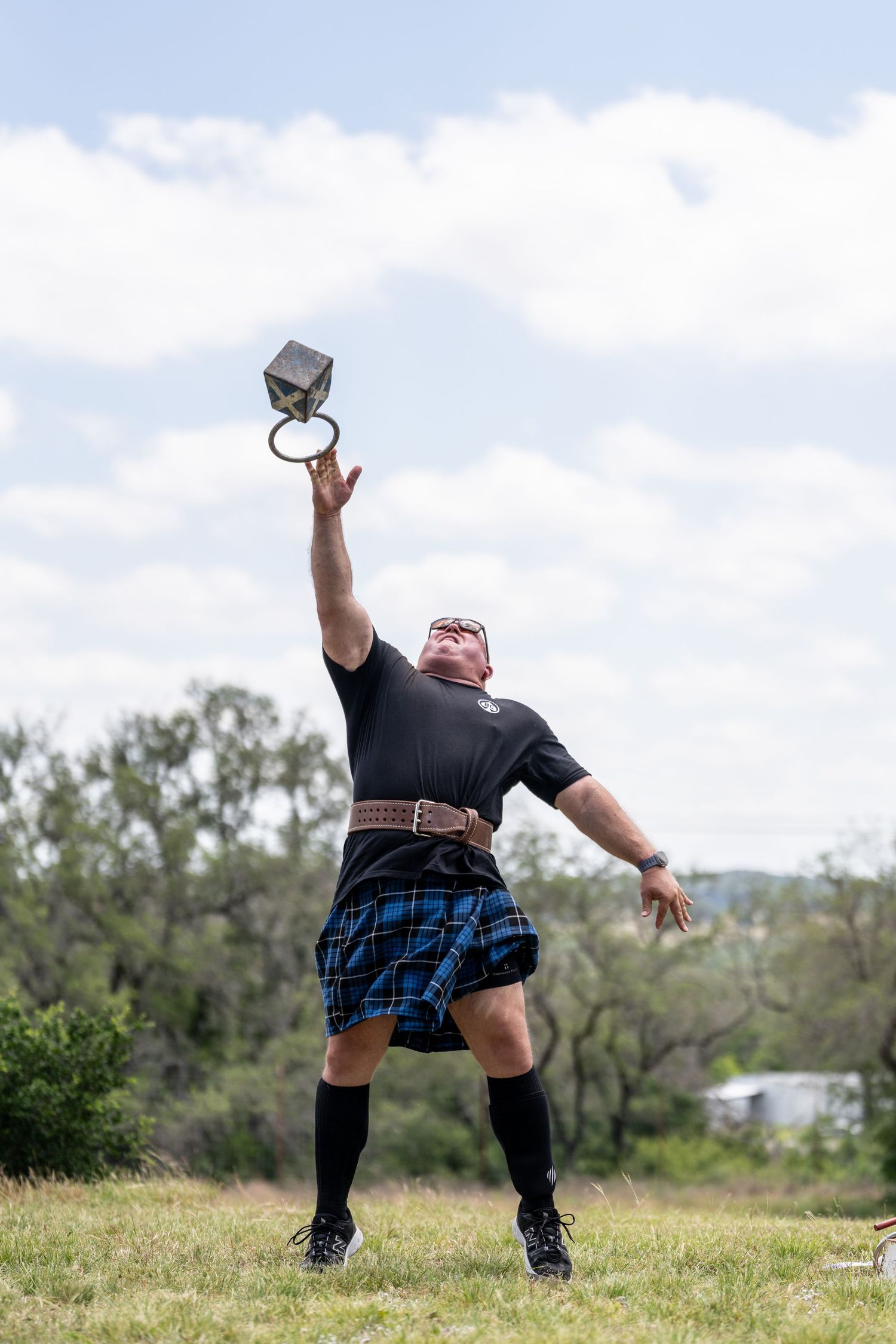 Man in blue tartan kilt performs Highland Games weight toss, reaching arm skyward on grassy field.