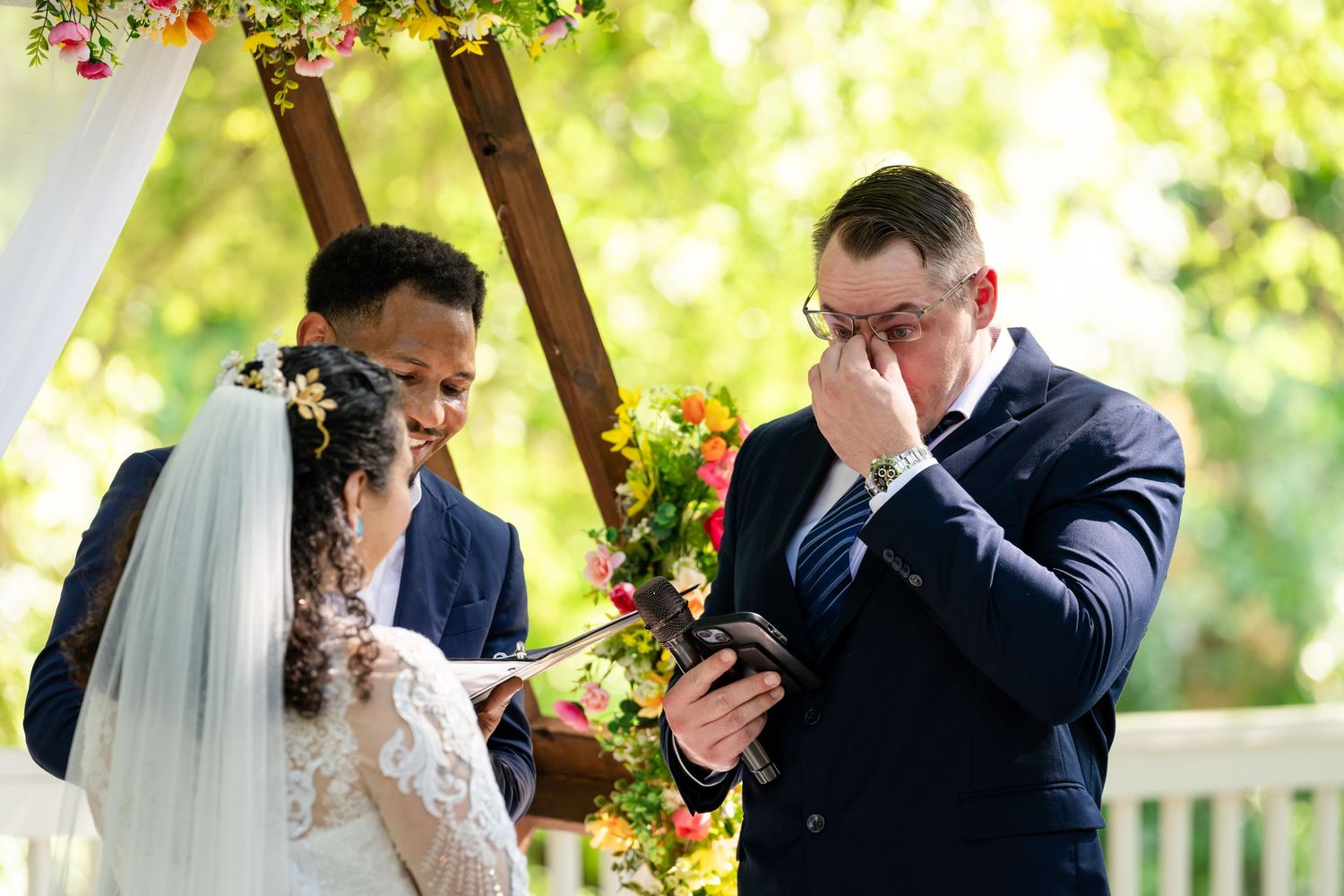 Emotional groom wiping tears during outdoor wedding ceremony while bride smiles, standing beneath floral arch.