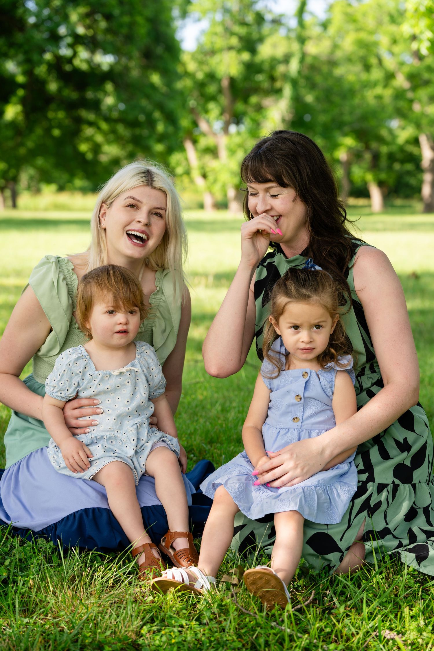 Two women and two toddler girls sitting on green grass in a sunny park, smiling and enjoying a warm summer day.