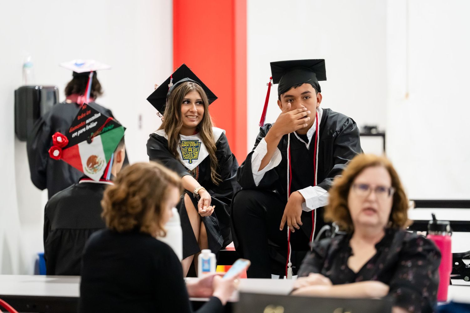 Graduates in caps and gowns chat at a registration table during a college graduation ceremony.