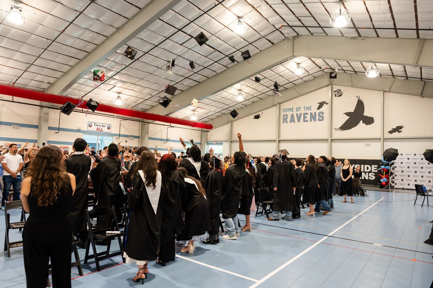 Graduates in black gowns toss caps at an indoor Ravens school graduation ceremony in a gymnasium.