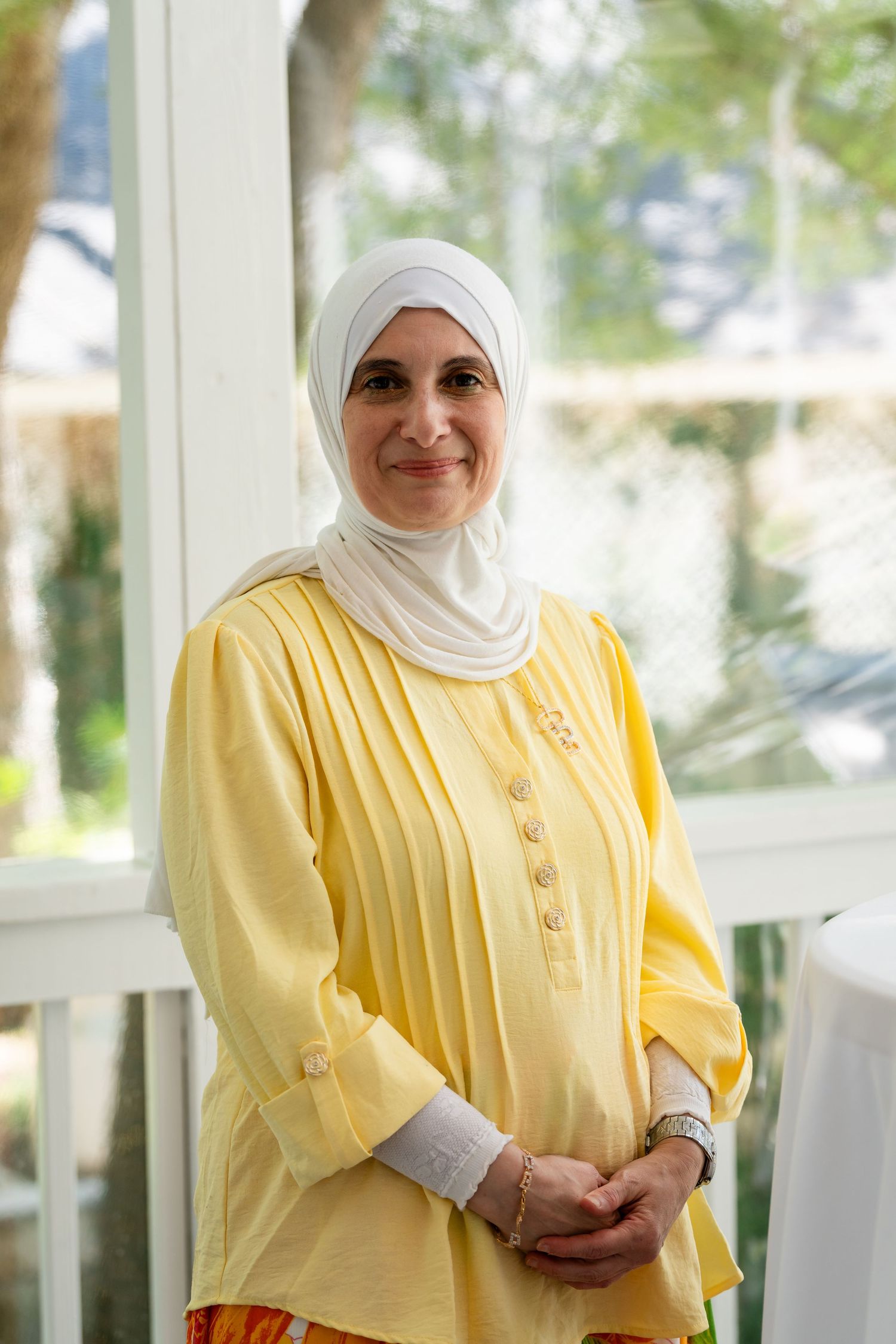 Smiling woman wearing a white hijab and yellow tunic top stands indoors near a bright window.