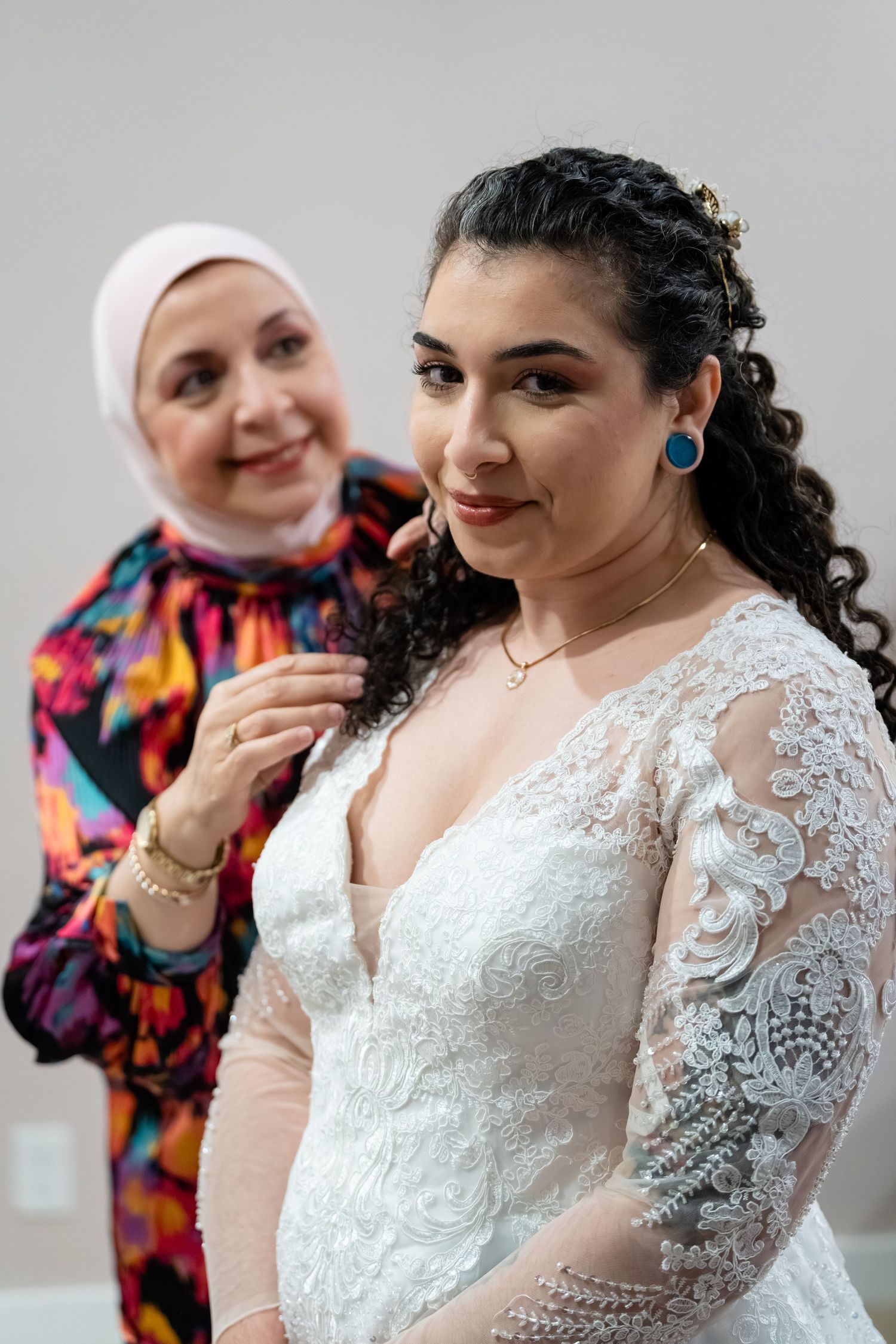 A bride in a white lace gown gets ready as an older woman in a hijab and colorful dress adjusts her necklace.