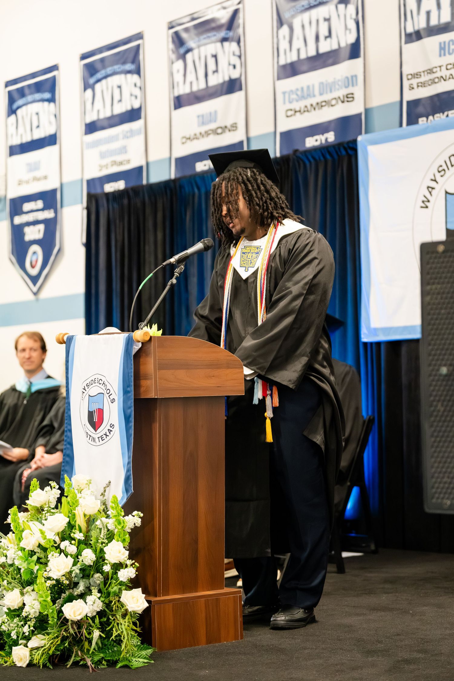 Graduate in cap and gown delivers speech at podium during high school graduation ceremony in school gymnasium.