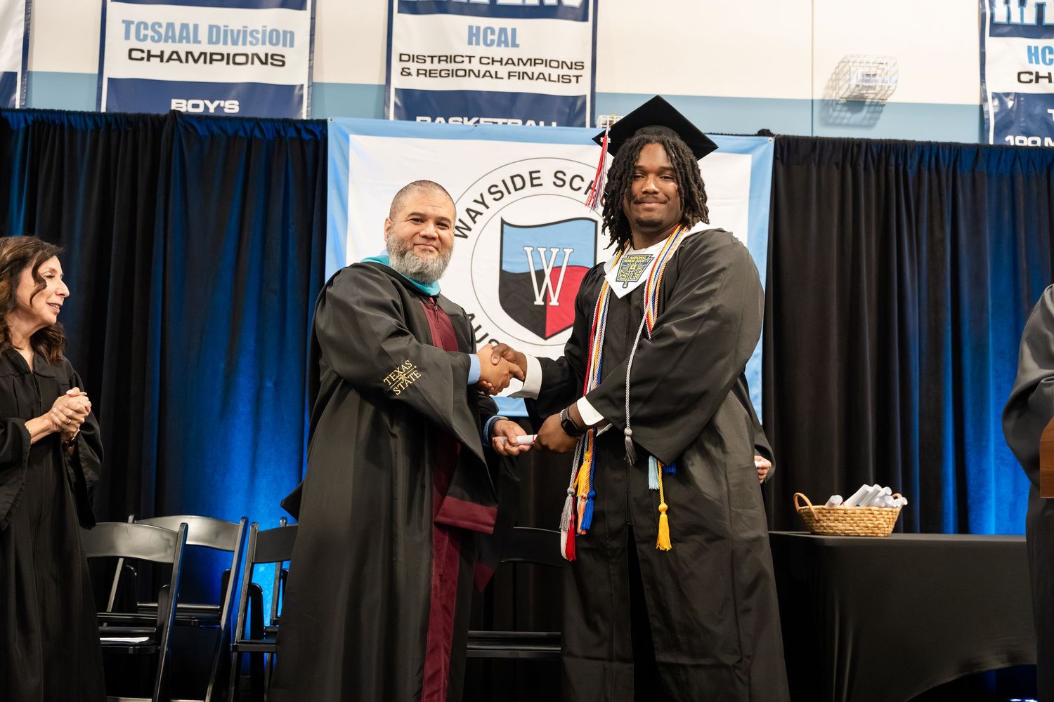 Graduate shaking hands with administrator in academic regalia at Bayside School graduation ceremony.